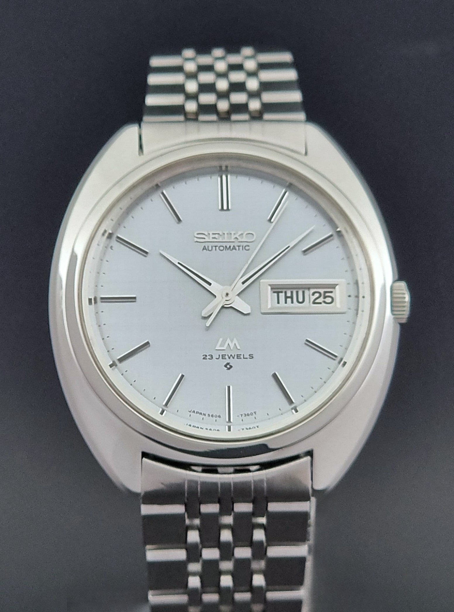 Vintage Seiko Lord Matic 5606-7150 stainless steel wristwatch with a metallic band on a dark gray background front view