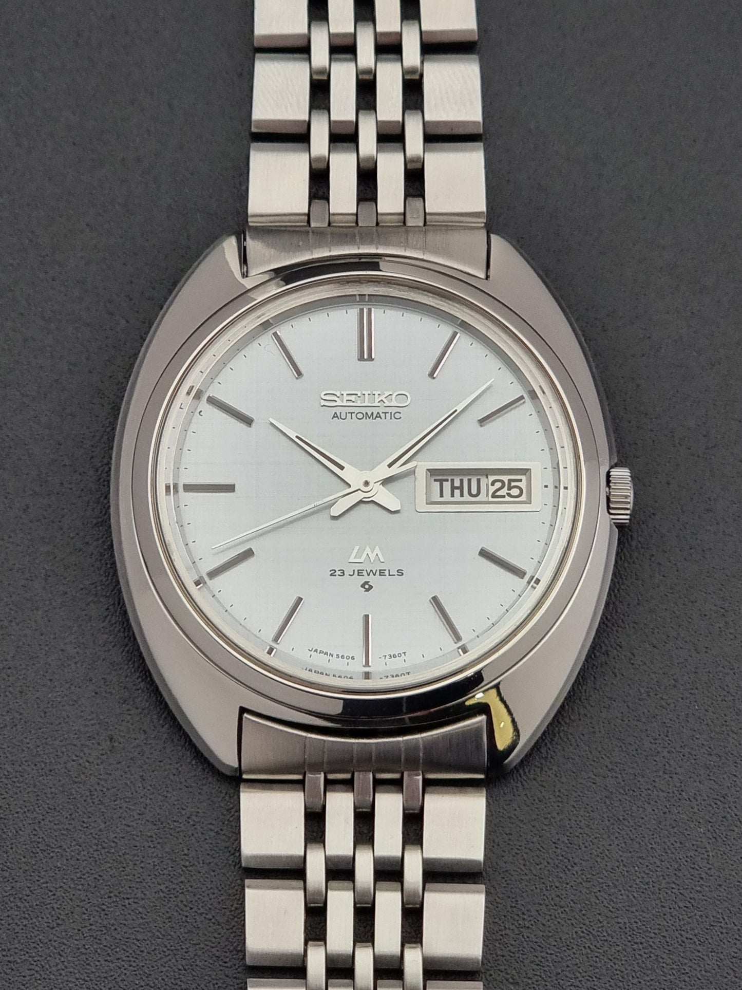 Vintage Seiko Lord Matic 5606-7150 stainless steel wristwatch with a metallic band on a dark gray background front