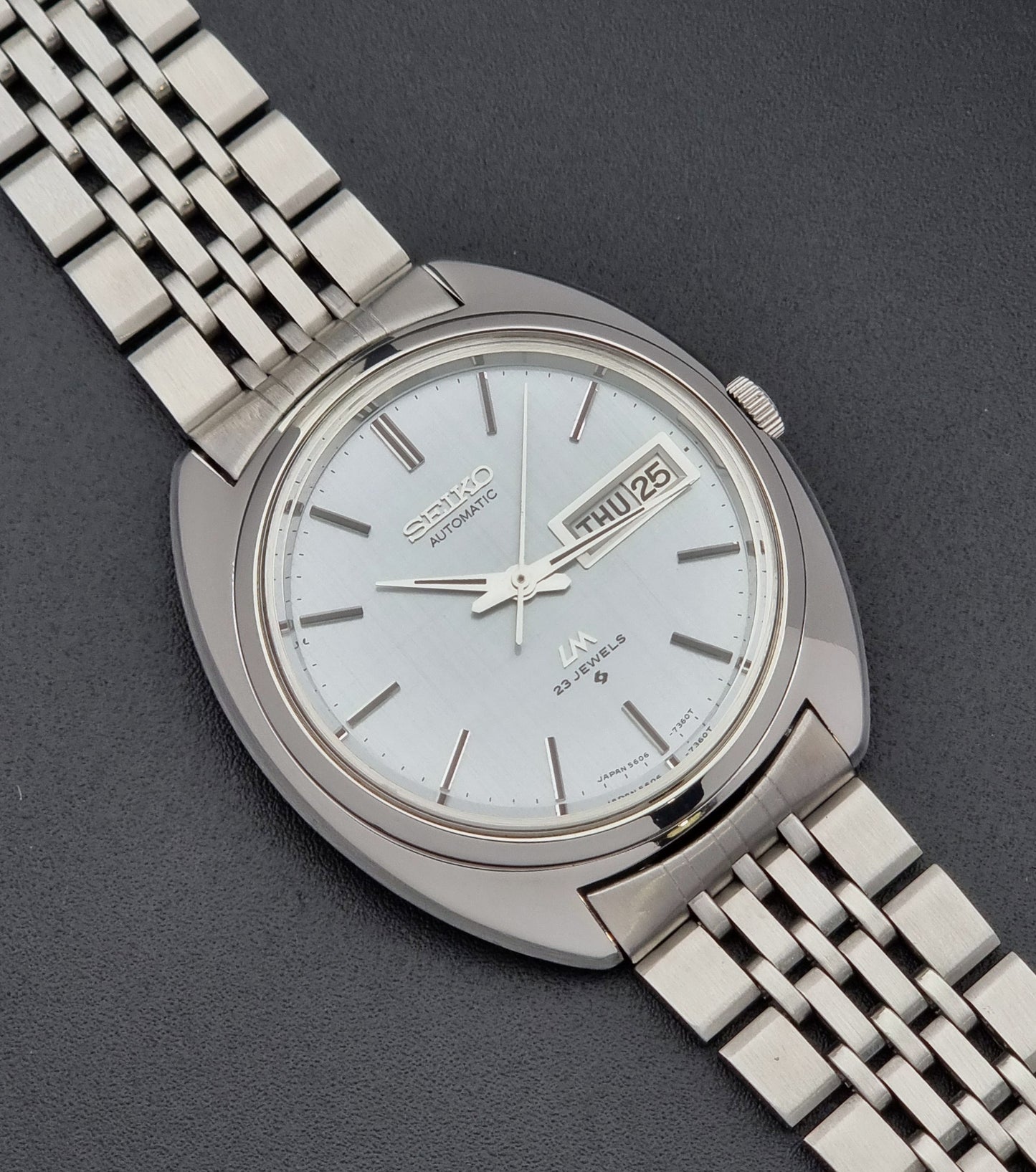 Vintage Seiko Lord Matic 5606-7150 stainless steel wristwatch with a metallic band on a dark gray background dial