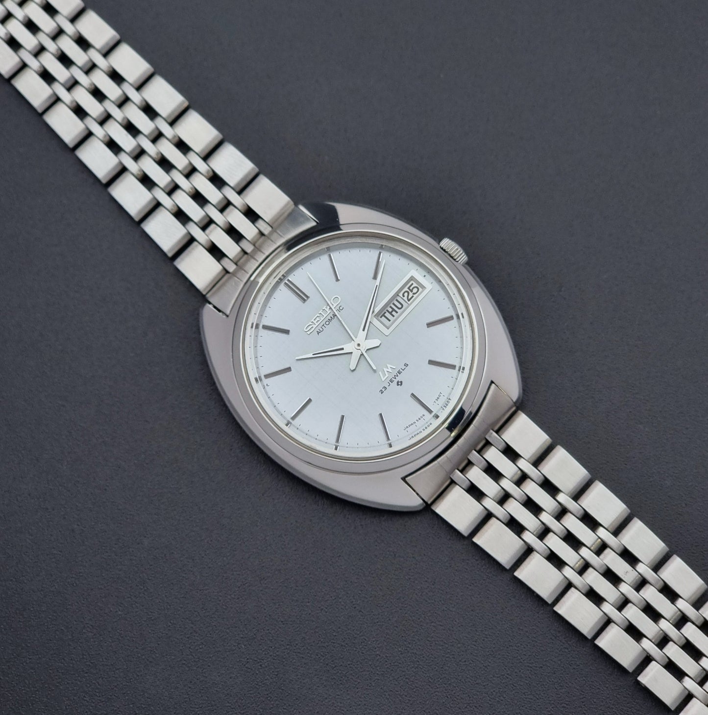 Vintage Seiko Lord Matic 5606-7150 stainless steel wristwatch with a metallic band on a dark gray background
