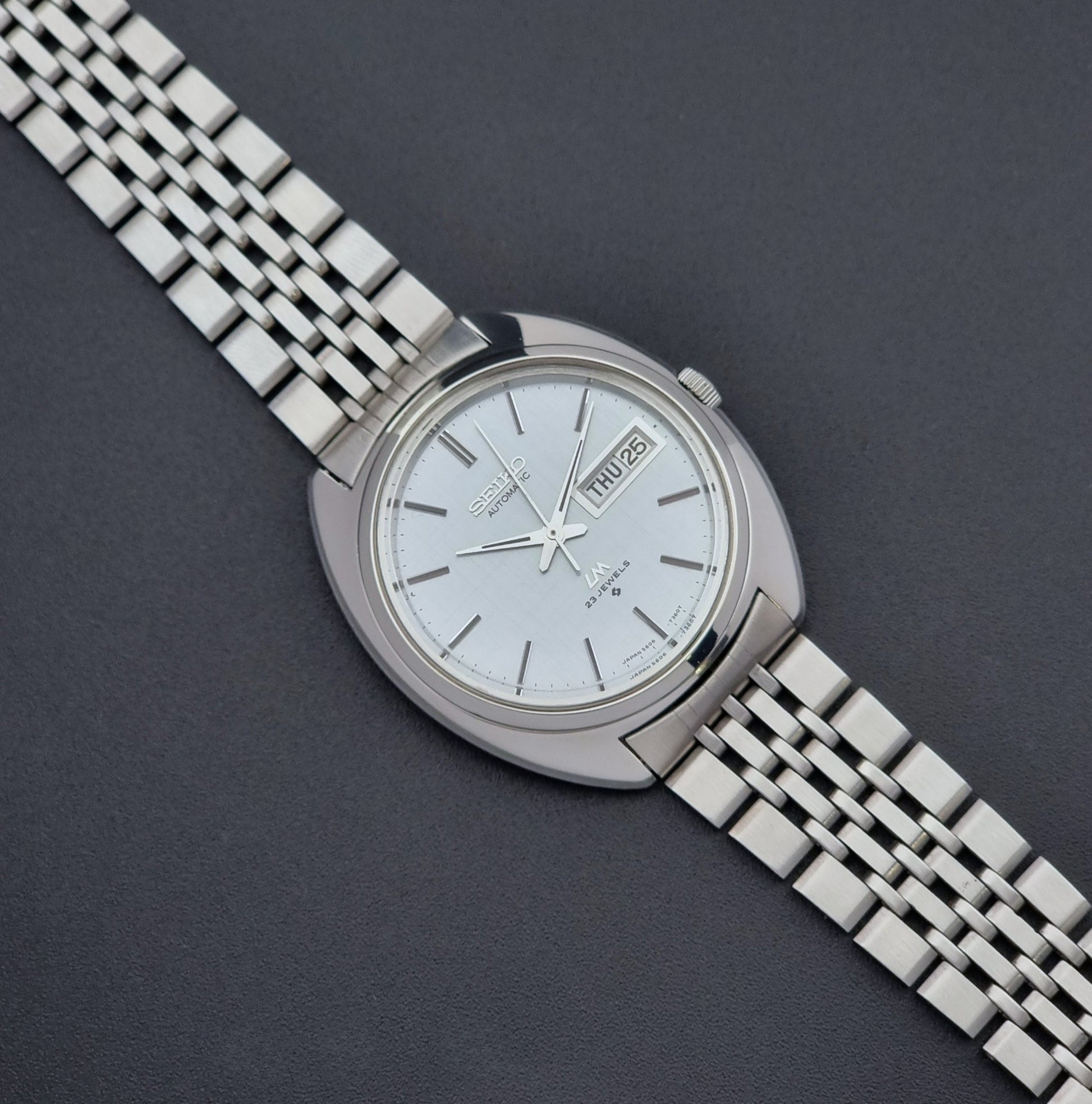 Vintage Seiko Lord Matic 5606-7150 stainless steel wristwatch with a metallic band on a dark gray background