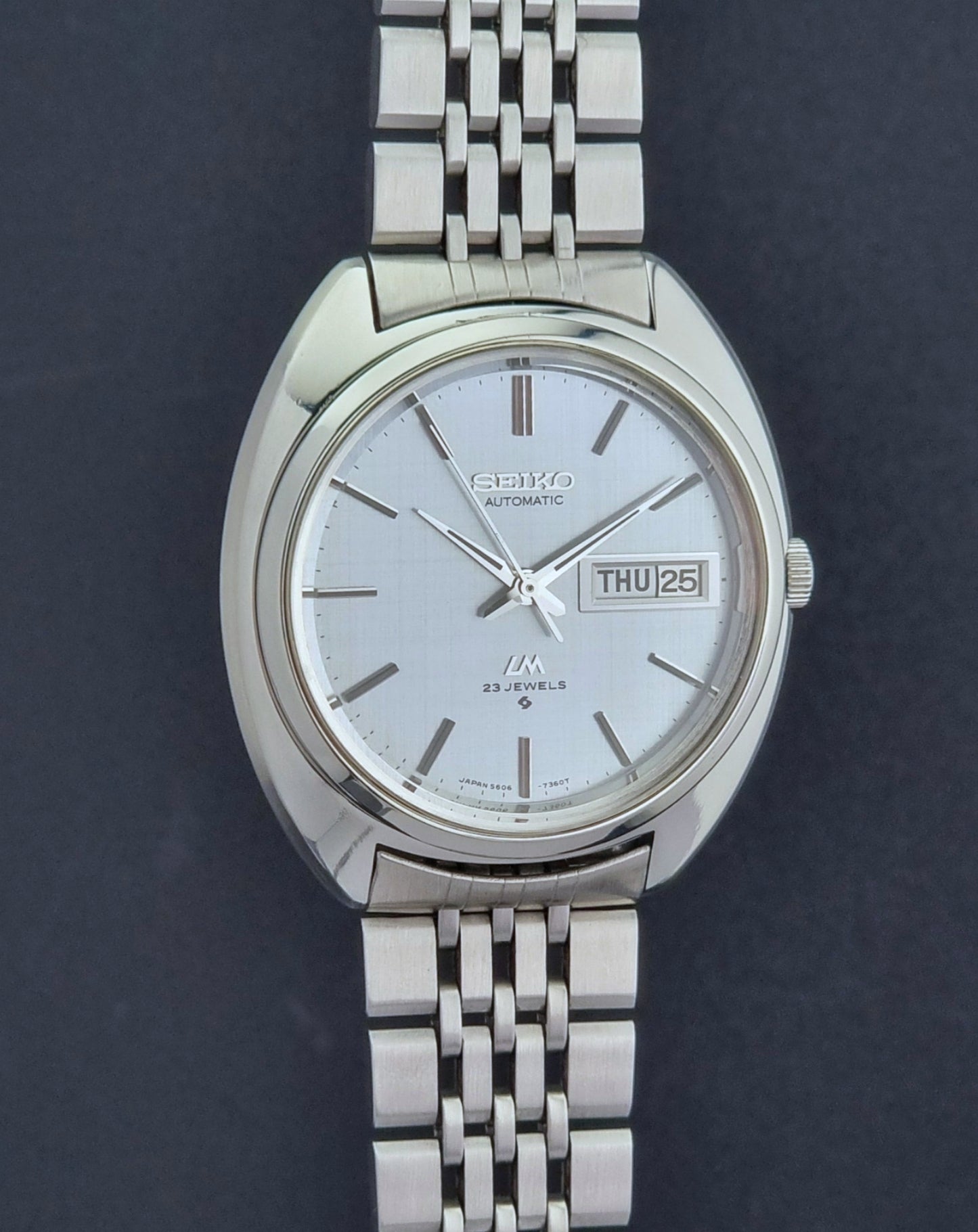 Vintage Seiko Lord Matic 5606-7150 stainless steel wristwatch with a metallic band on a dark gray background side profile