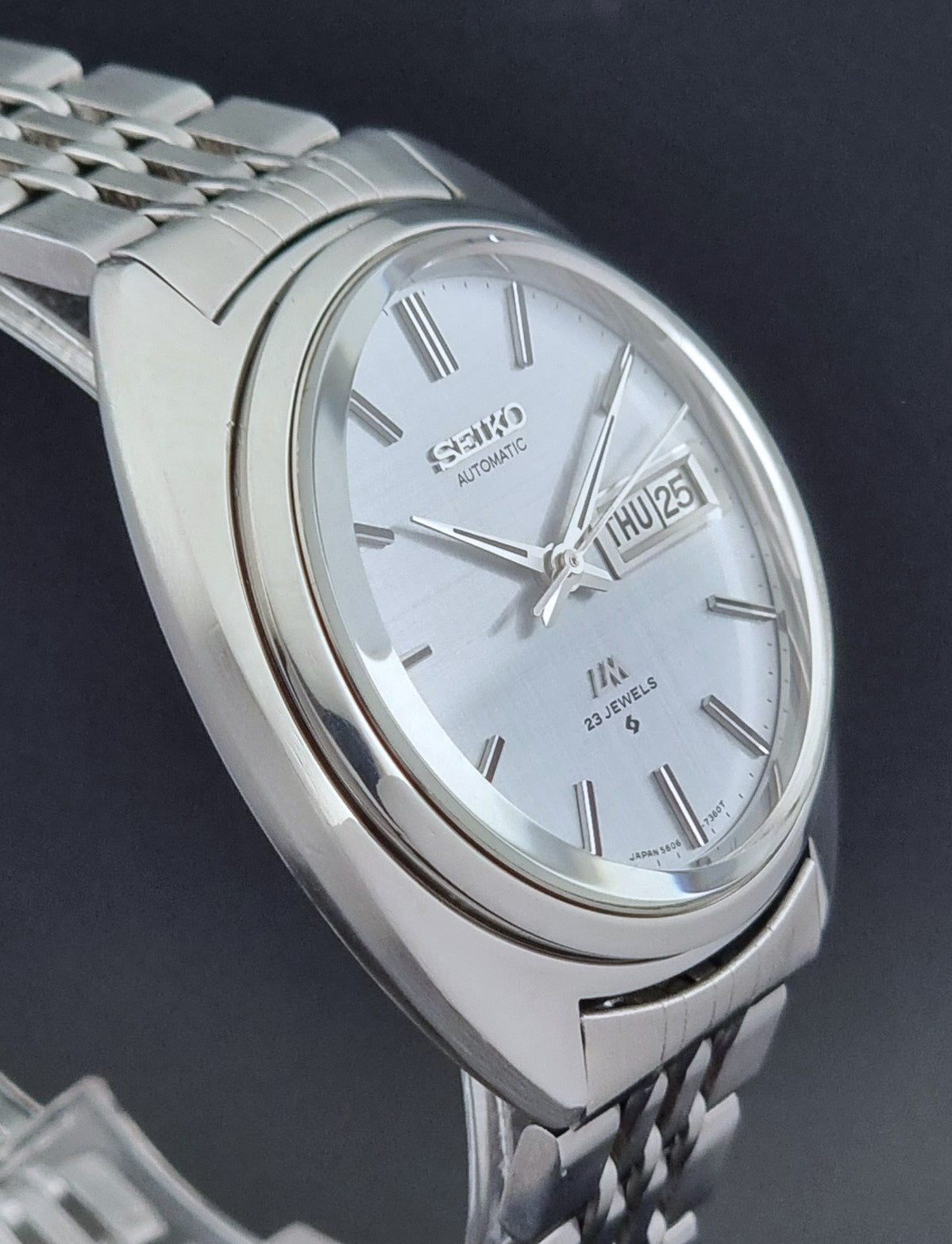 Vintage Seiko Lord Matic 5606-7150 stainless steel wristwatch with a metallic band on a dark gray background side