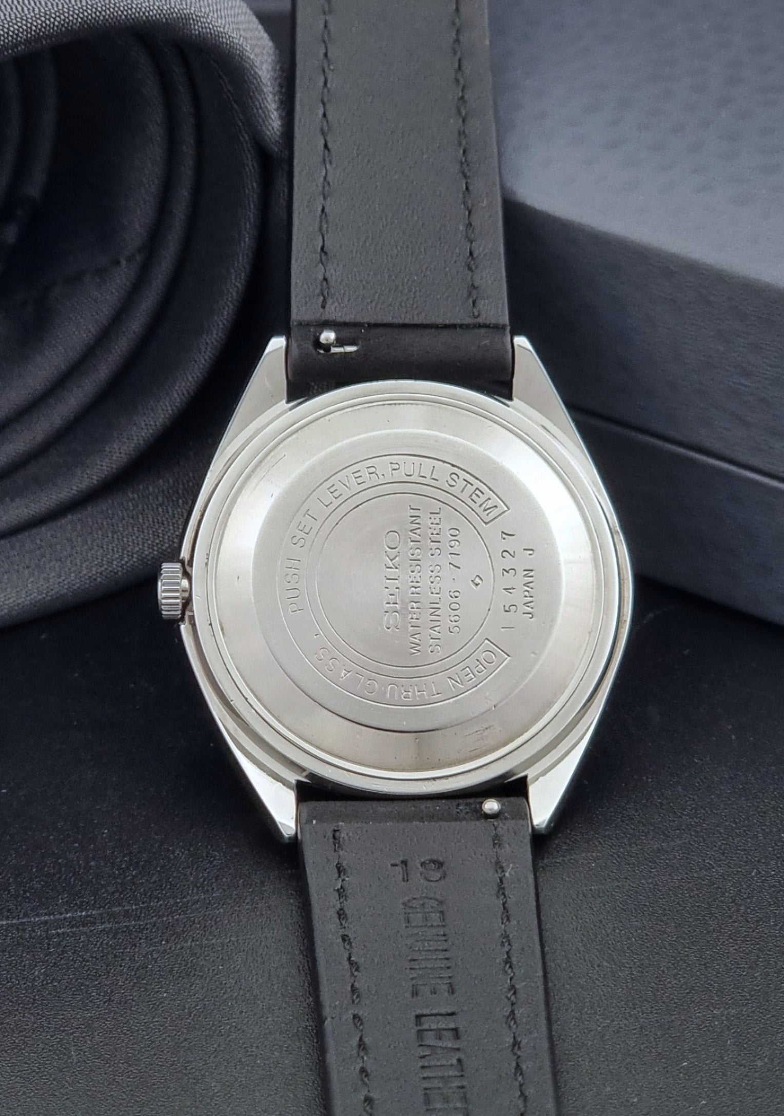 Vintage Seiko Wristwatch LordMatic 5606-7190 black strap and silver case on a dark gray background case back view