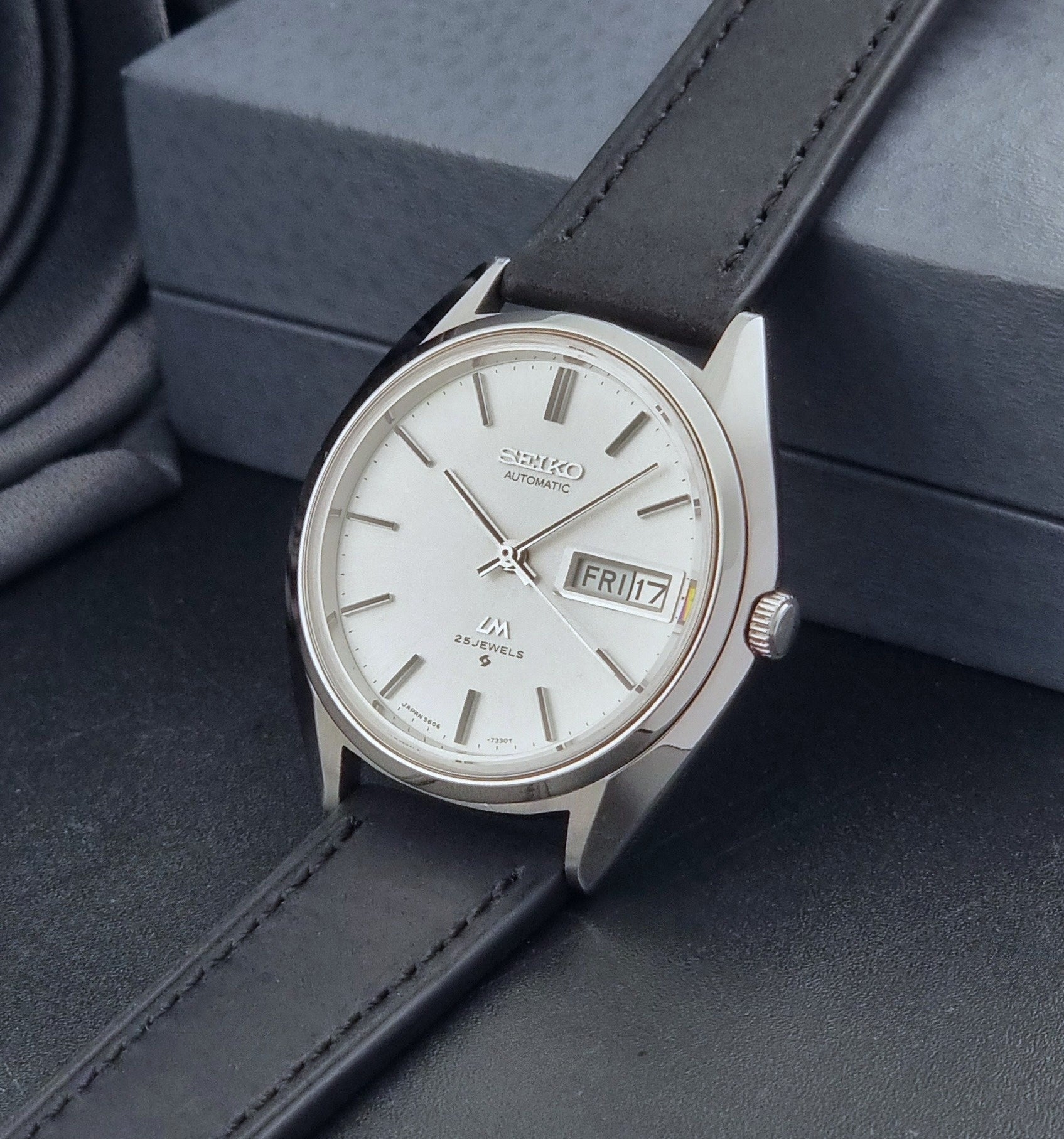 Vintage Seiko Wristwatch LordMatic 5606-7190 black strap and silver case on a dark gray background crown view