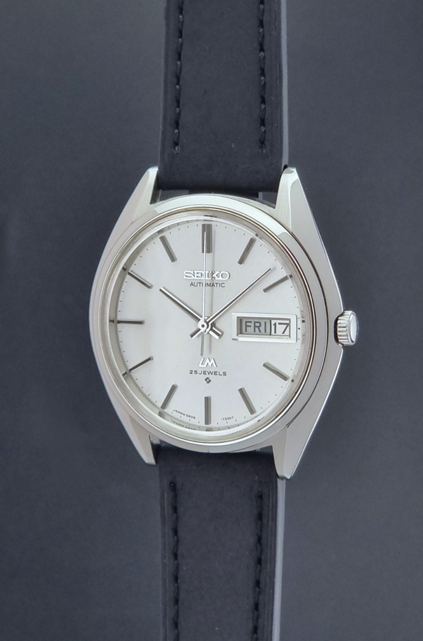 Vintage Seiko Wristwatch LordMatic 5606-7190 black strap and silver case on a dark gray background crown profile view