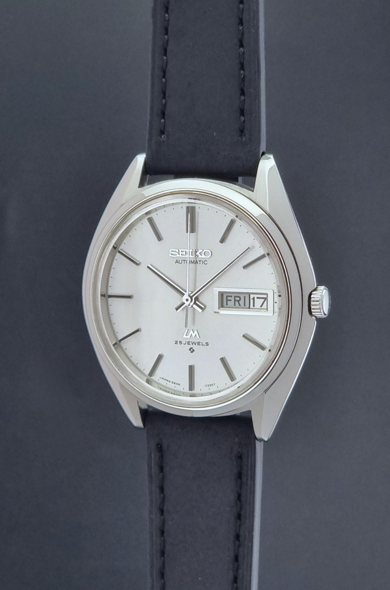 Vintage Seiko Wristwatch LordMatic 5606-7190 black strap and silver case on a dark gray background crown profile view