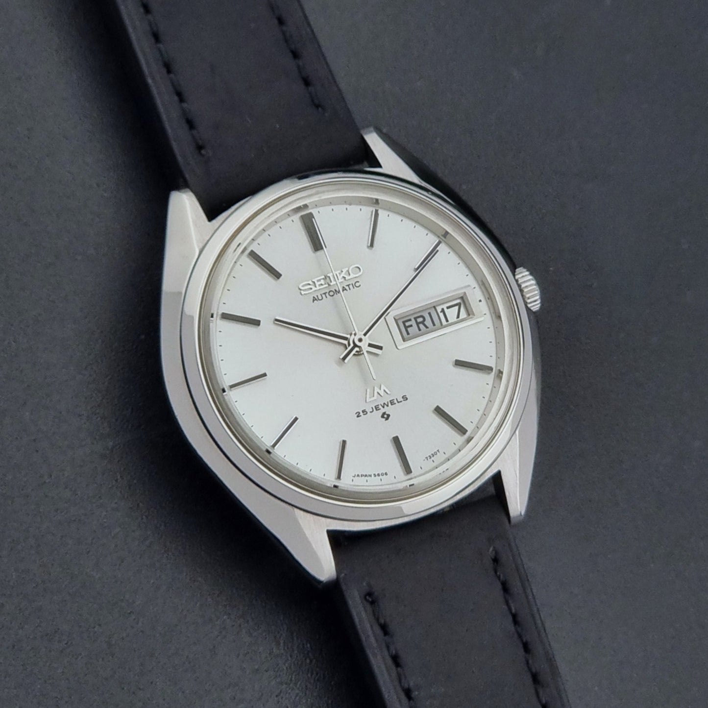 Vintage Seiko Wristwatch LordMatic 5606-7190 black strap and silver case on a dark gray background front dial view