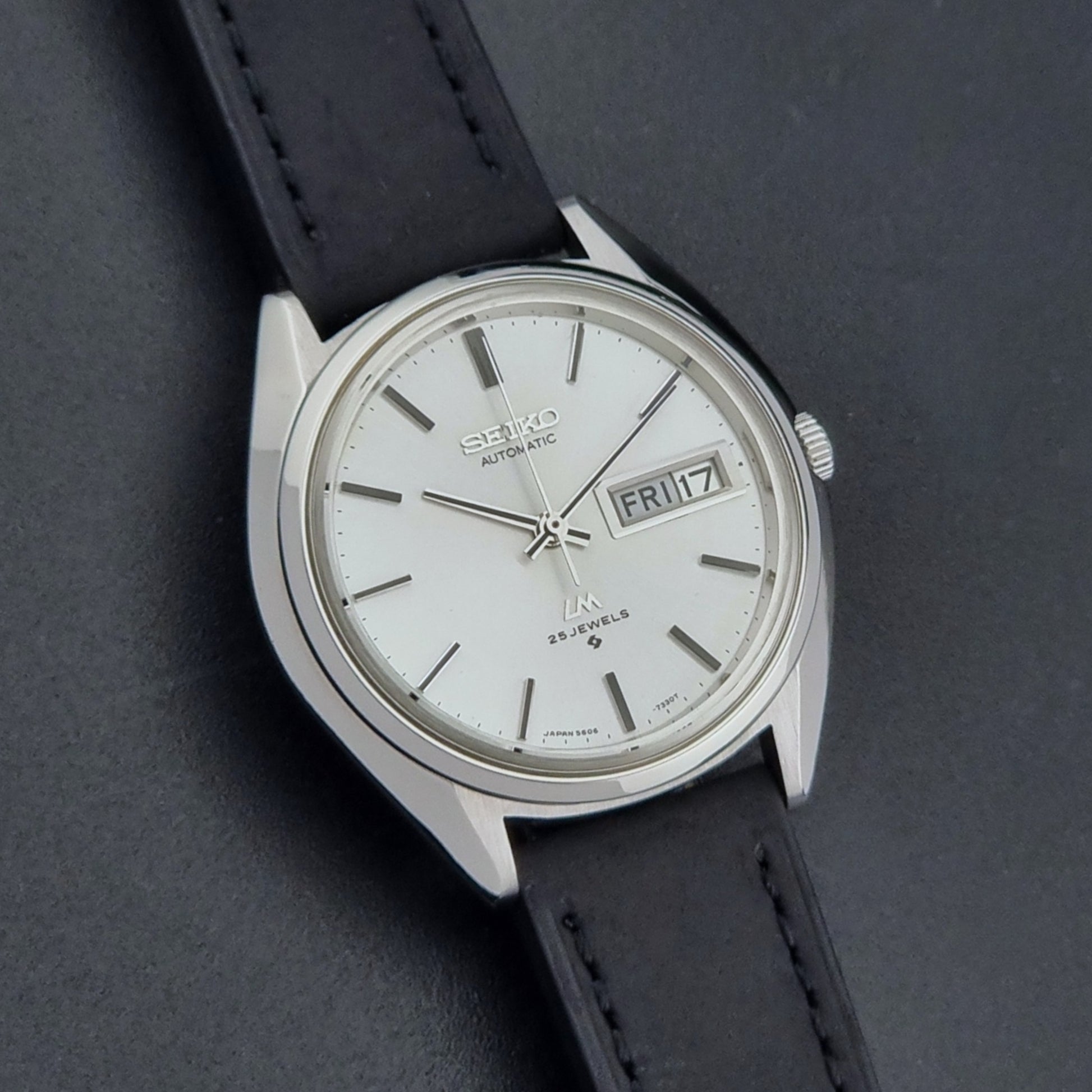 Vintage Seiko Wristwatch LordMatic 5606-7190 black strap and silver case on a dark gray background front dial view