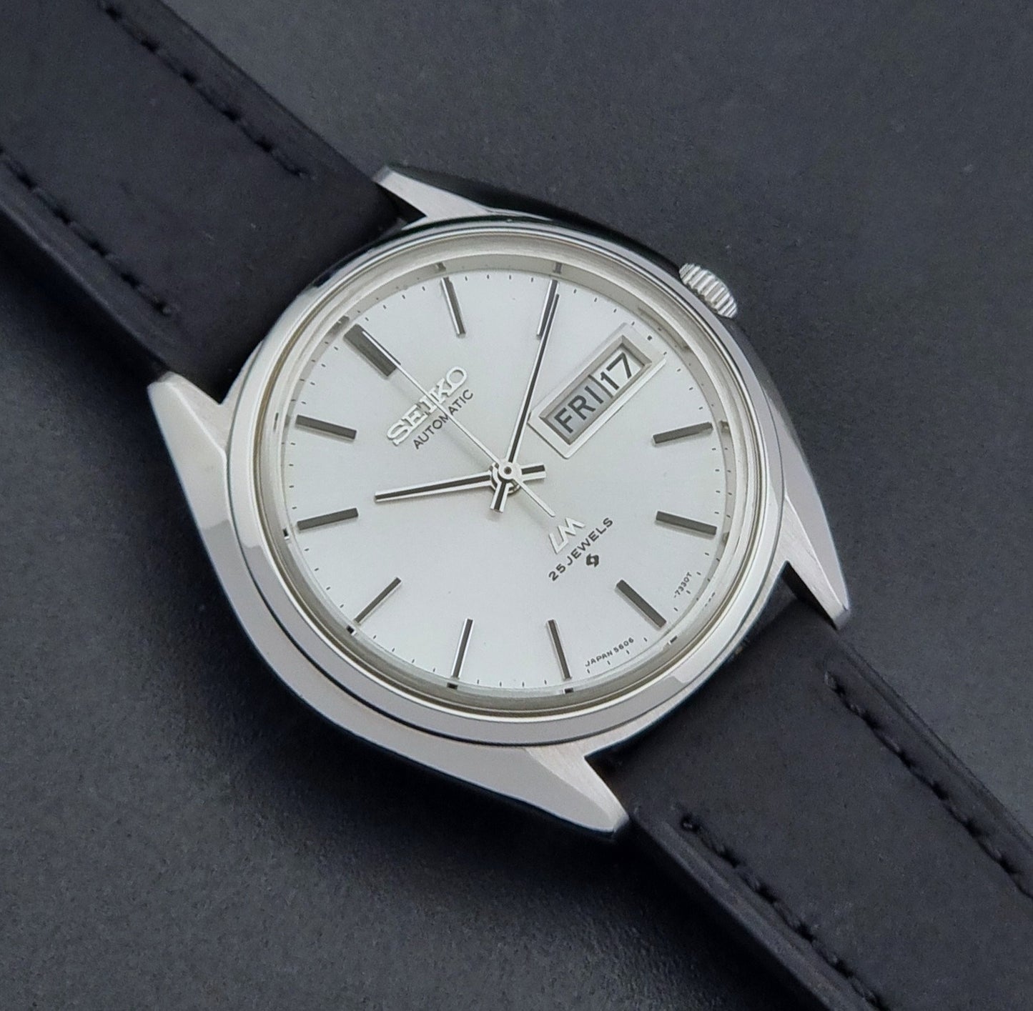 Vintage Seiko Wristwatch LordMatic 5606-7190 black strap and silver case on a dark gray background close front view