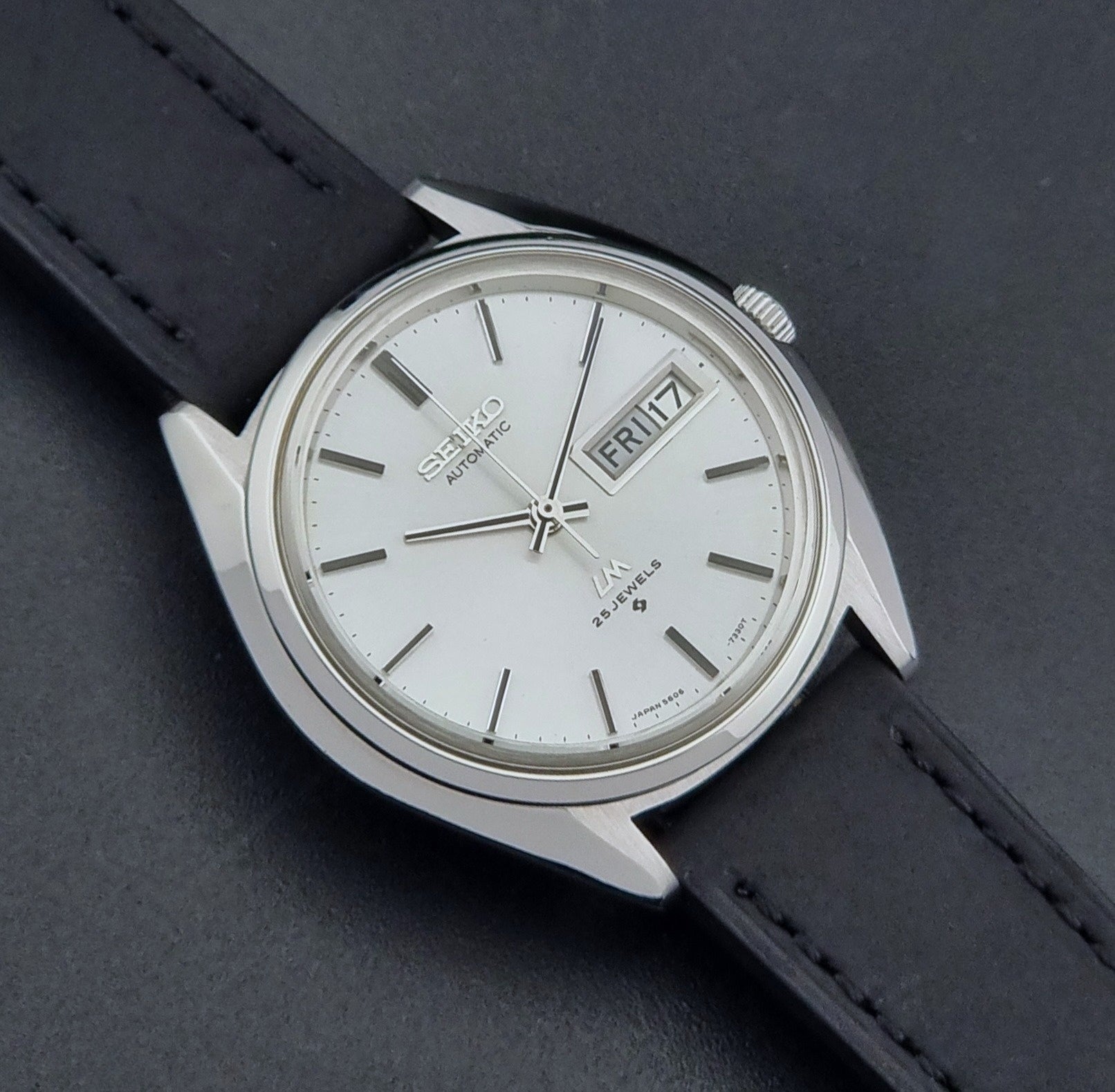 Vintage Seiko Wristwatch LordMatic 5606-7190 black strap and silver case on a dark gray background close front view
