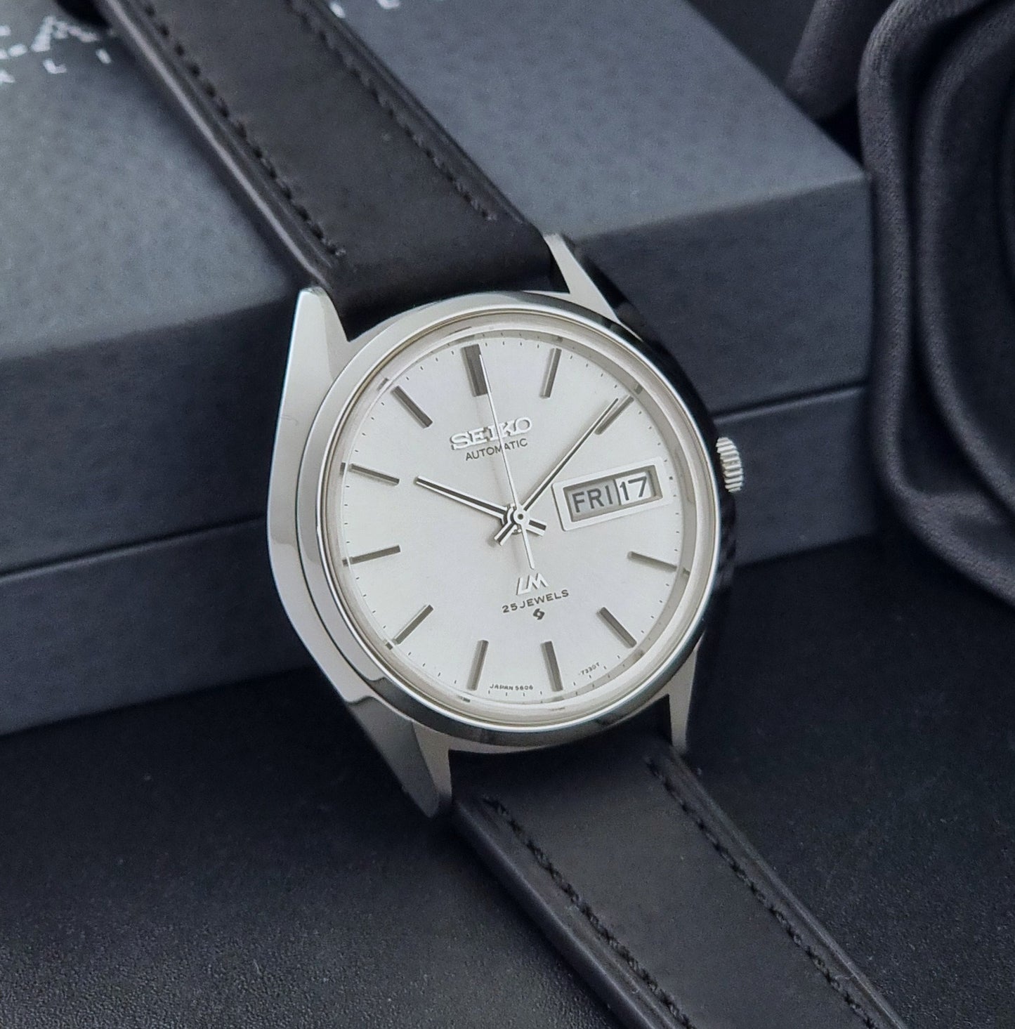 Vintage Seiko Wristwatch LordMatic 5606-7190 black strap and silver case on a dark gray background side view