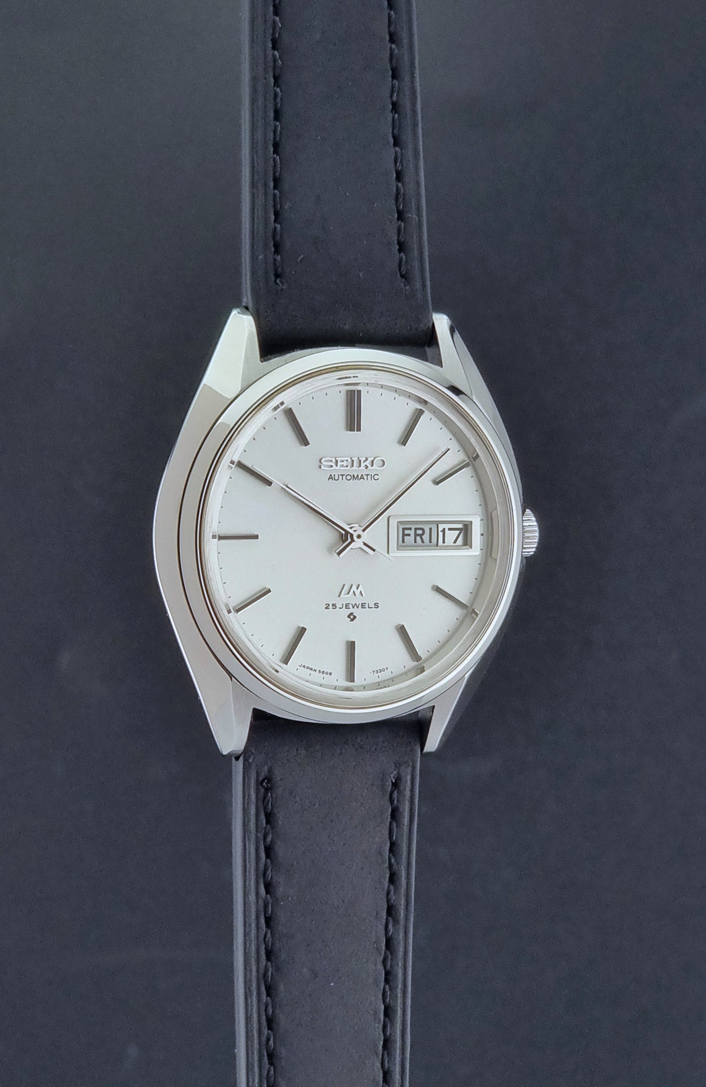 Vintage Seiko Wristwatch LordMatic 5606-7190 black strap and silver case on a dark gray background side profile view