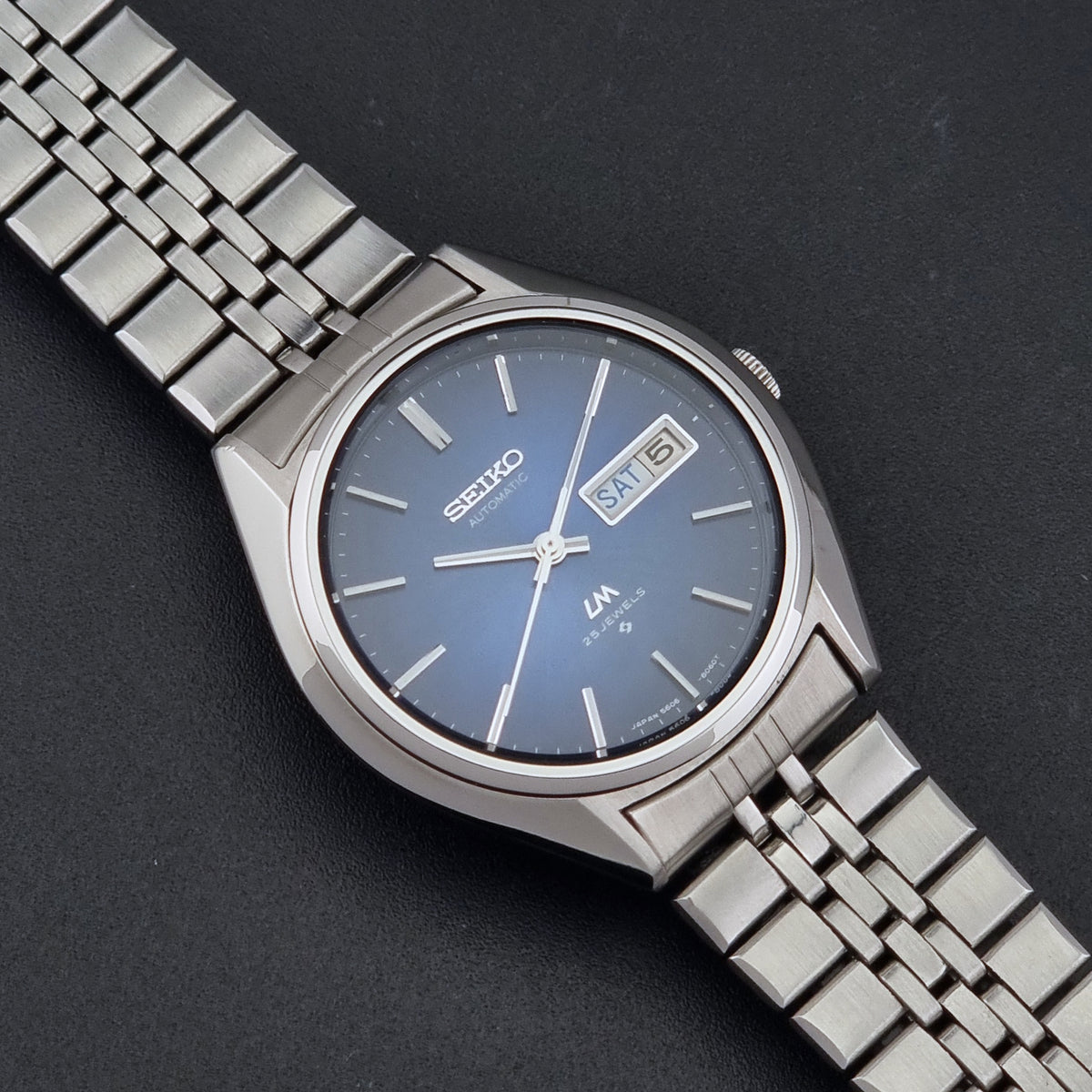 A Vintage Seiko watch LordMatic 5606-8031 with a silver stainless steel case and link bracelet, featuring a blue sunburst dial with silver-tone hour markers and hands, and a day-date window at 3 o'clock, shown on a dark textured background.