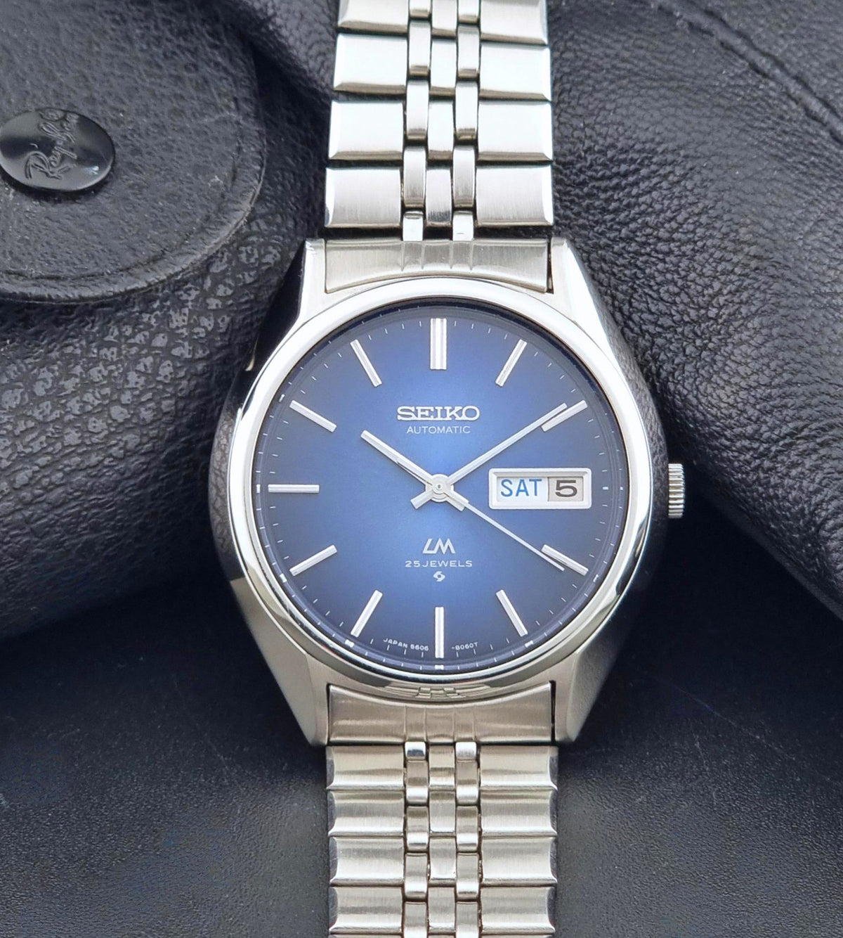 A Vintage Seiko watch LordMatic 5606-8031 Automatic watch with a blue dial and a silver stainless steel link bracelet, resting on a textured black leather surface.