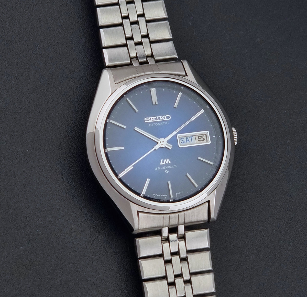 A Vintage Seiko watch LordMatic 5606-8031 with a silver stainless steel case and link bracelet, featuring a blue dial with silver indices and a day-date window at 3 o'clock, set against a dark textured background.