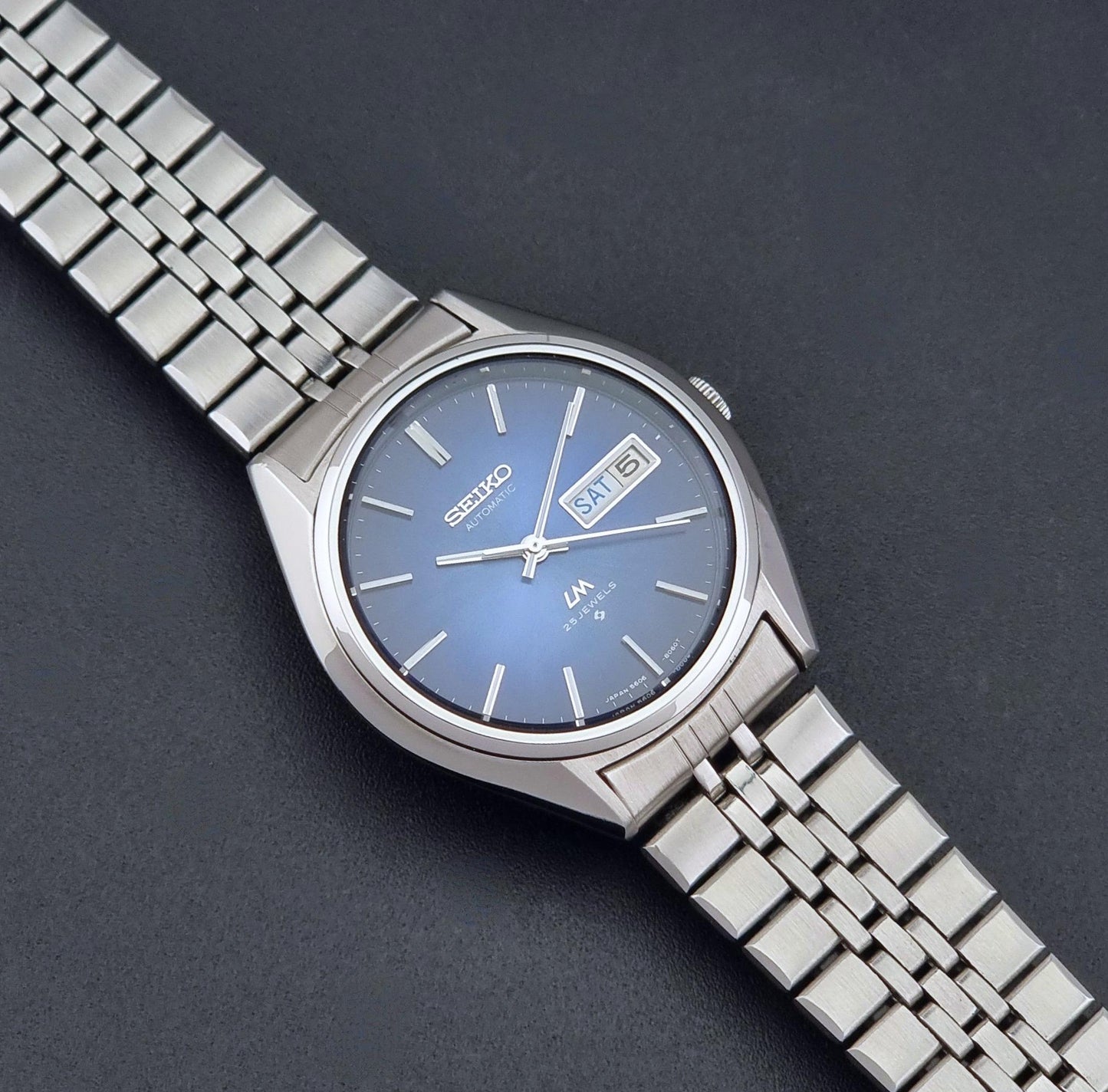A Vintage Seiko watch LordMatic 5606-8031 with a silver stainless steel link bracelet and a blue sunburst dial, shown diagonally on a dark gray textured background.