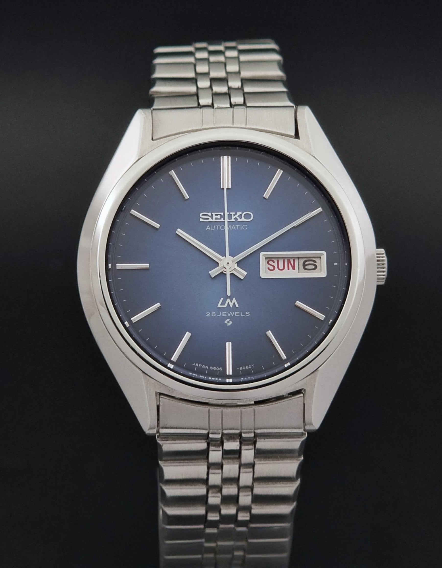 A Vintage Seiko watch LordMatic 5606-8031 with a silver stainless steel case and metal bracelet, featuring a blue dial with silver-tone hour markers and a day-date window showing 'SUN 6'.