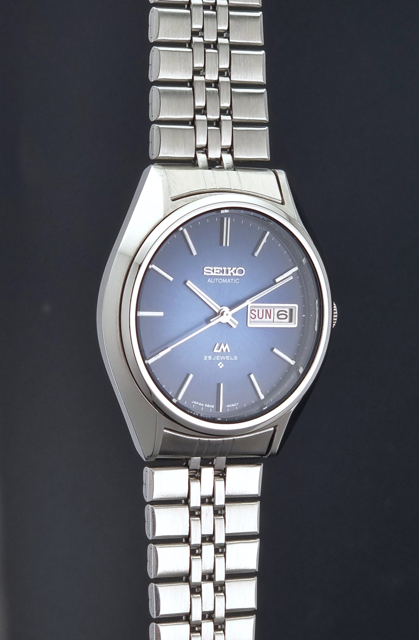 A Vintage Seiko watch LordMatic 5606-8031 analog watch with a silver stainless steel link bracelet, a silver-toned case, and a blue dial featuring a day and date window.