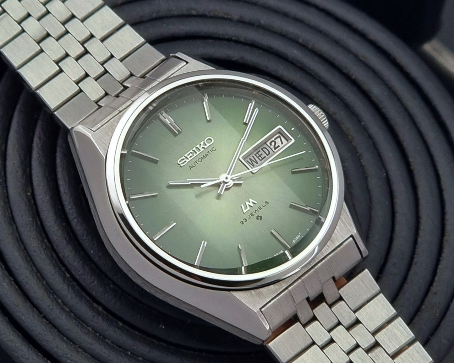 Vintage Seiko Lord Matic 56206-8130 watch with green face and silver metal strap on a dark textured background close up dial