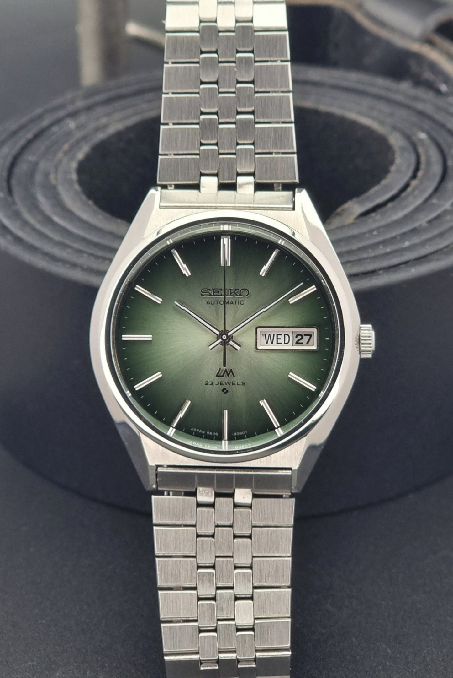 Vintage Seiko Lord Matic 56206-8130 watch with green face and silver metal strap on a dark textured background front view