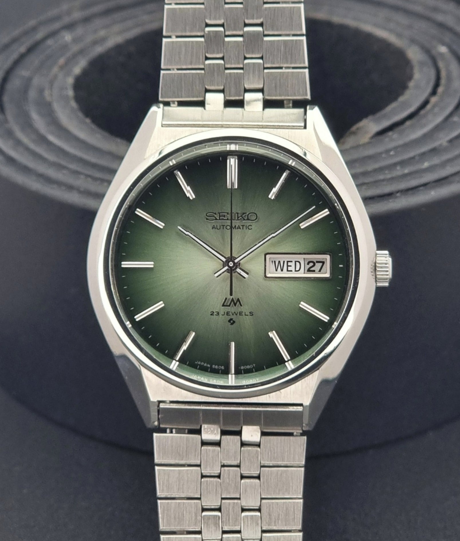 Vintage Seiko Lord Matic 56206-8130 watch with green face and silver metal strap on a dark textured background close up dial view
