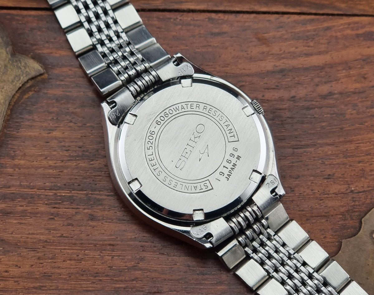 Close-up of the back of a stainless steel wristwatch with a metal link bracelet, resting on a dark wood surface. The watch case and bracelet are silver-toned
