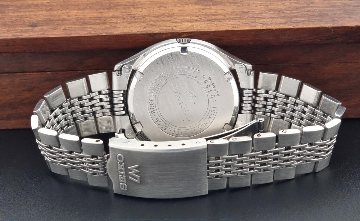 The back of a wristwatch with a brushed stainless steel case and a matching metal link bracelet, resting on a dark surface with a wooden background.