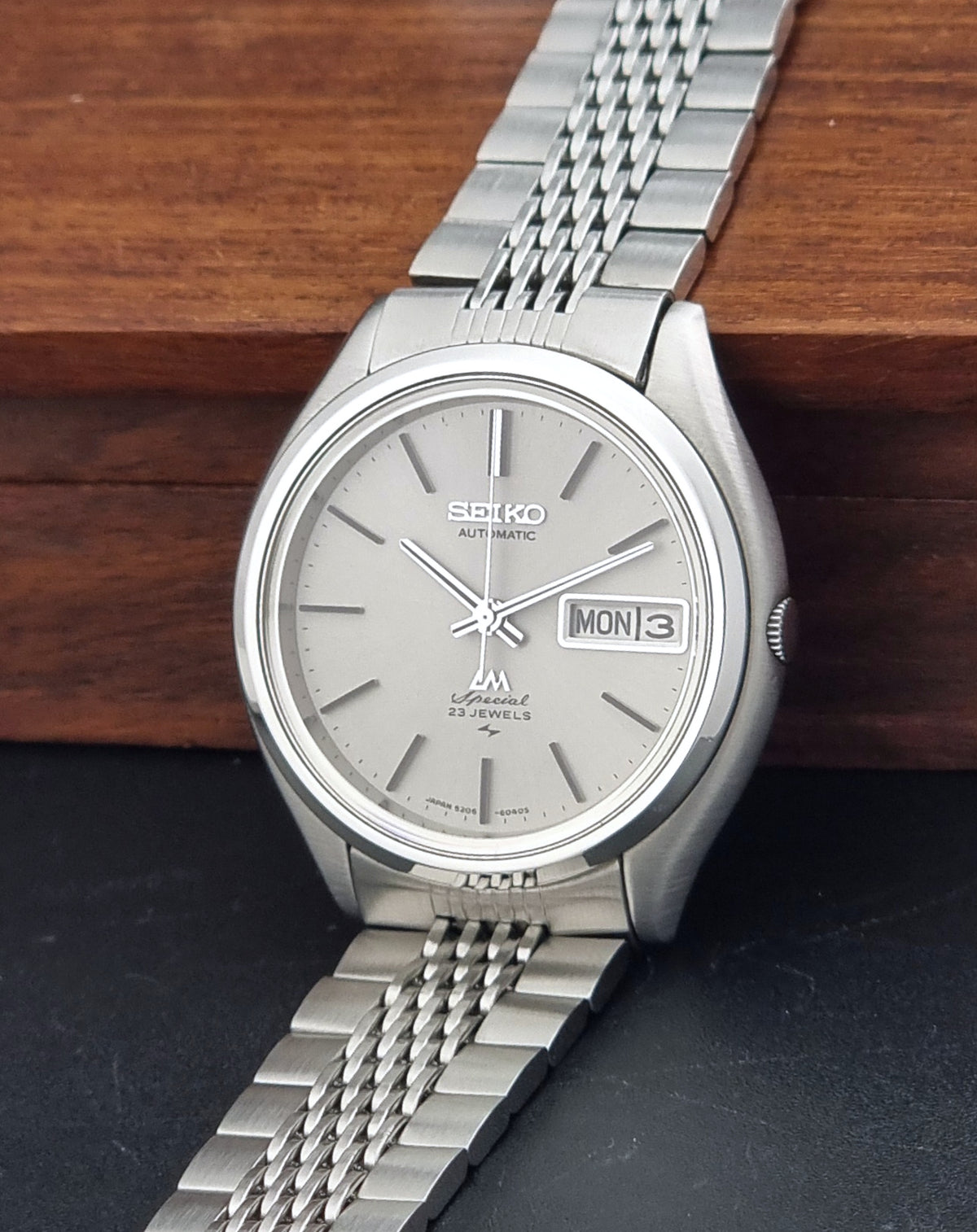 Vintage Seiko watch LordMatic Special 5206-6060 with a silver dial and matching metal link bracelet, resting on a dark surface with a wooden background.