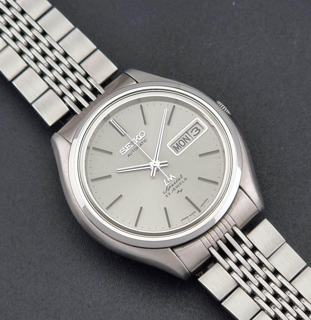 Vintage Seiko watch LordMatic Special 5206-6060 with a silver stainless steel case and a matching silver dial, featuring a date window at 3 o'clock and a three-link stainless steel bracelet, shown on a dark textured background.