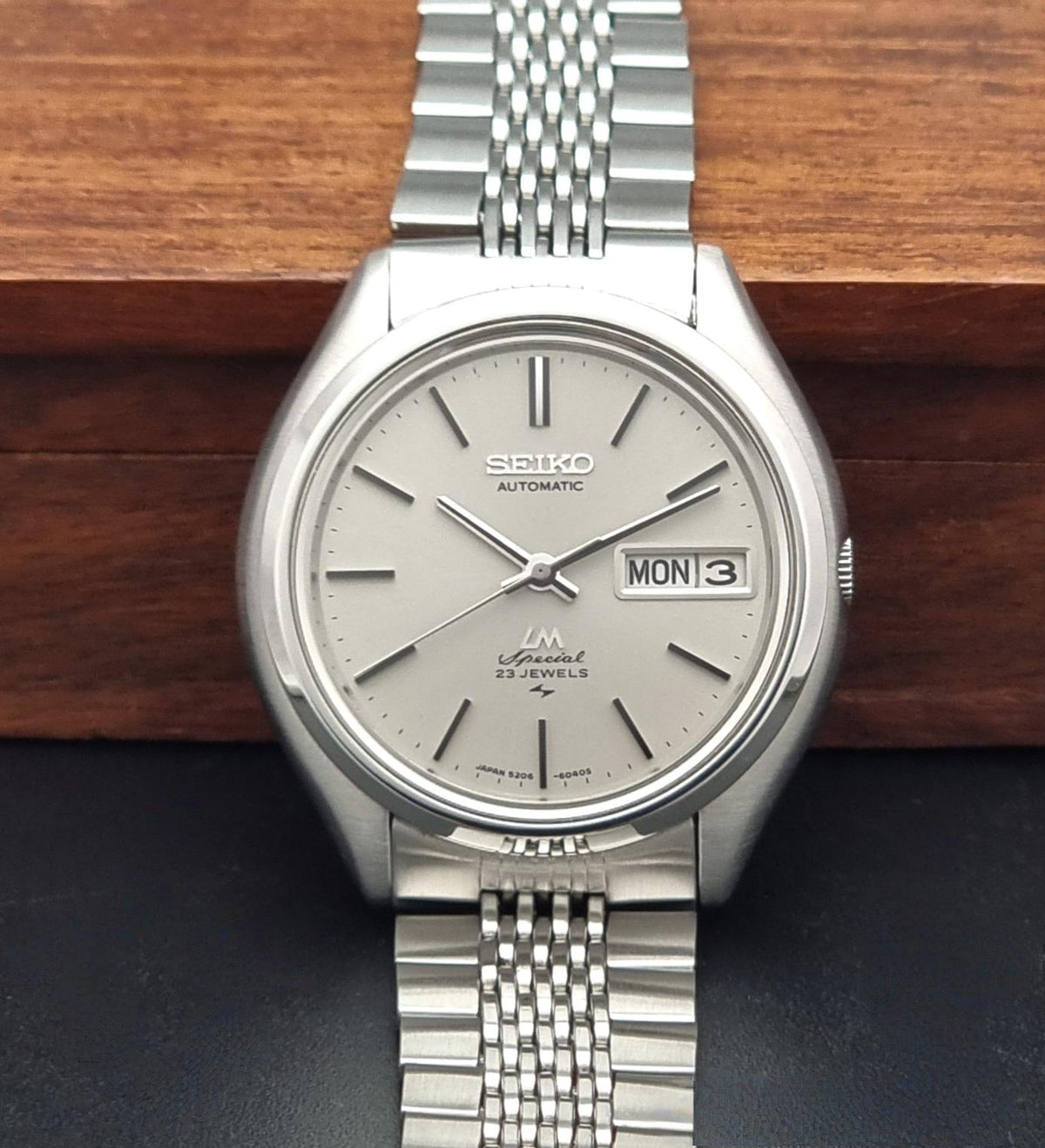 Vintage Seiko watch LordMatic Special 5206-6060 with a silver-toned stainless steel case and link bracelet, featuring a light gray dial with a day and date window, displayed on a dark surface with a wooden block in the background.