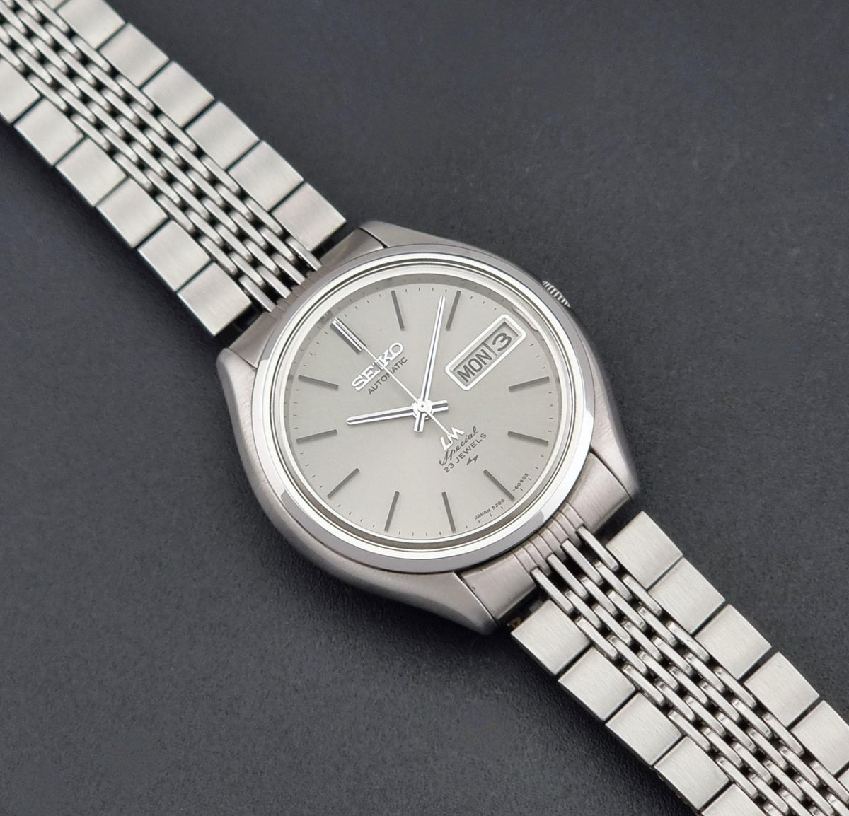 Vintage Seiko watch LordMatic Special 5206-6060 with a silver-toned three-link metal bracelet and a light gray dial, shown on a dark gray textured background.