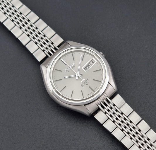 Vintage Seiko watch LordMatic Special 5206-6060 with a silver-toned three-link metal bracelet and a light gray dial, shown on a dark gray textured background.