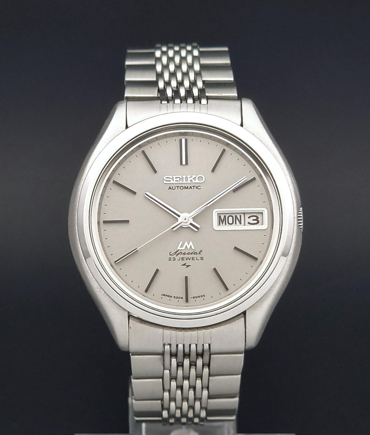 A Seiko LM Automatic 23 Jewels watch with a silver stainless steel case and link bracelet, featuring a light grey dial with stick indices and a day-date window.