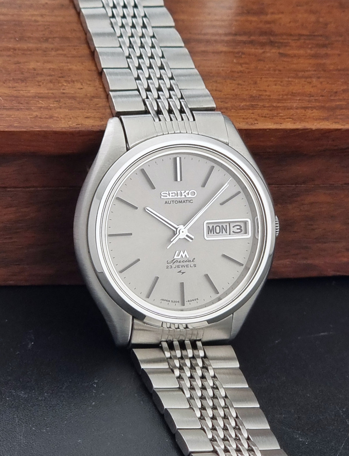 Vintage Seiko watch LordMatic Special 5206-6060 with a silver dial and stainless steel link bracelet, resting on a dark surface with a wooden block in the background.