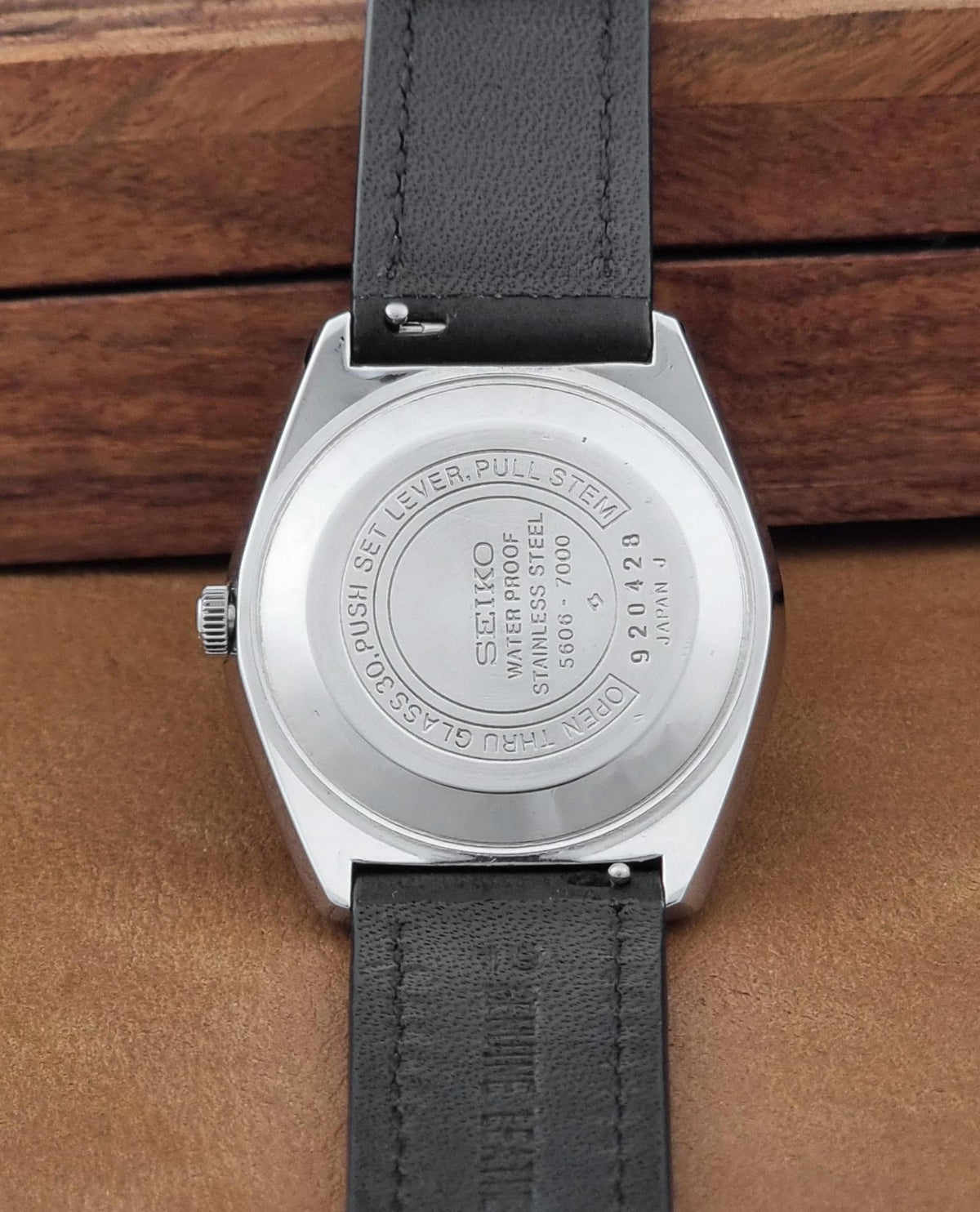 Back view of a Vintage Seiko watch LordMatic 5606-7000 with a silver stainless steel case and a black leather strap, resting on a brown leather surface.
