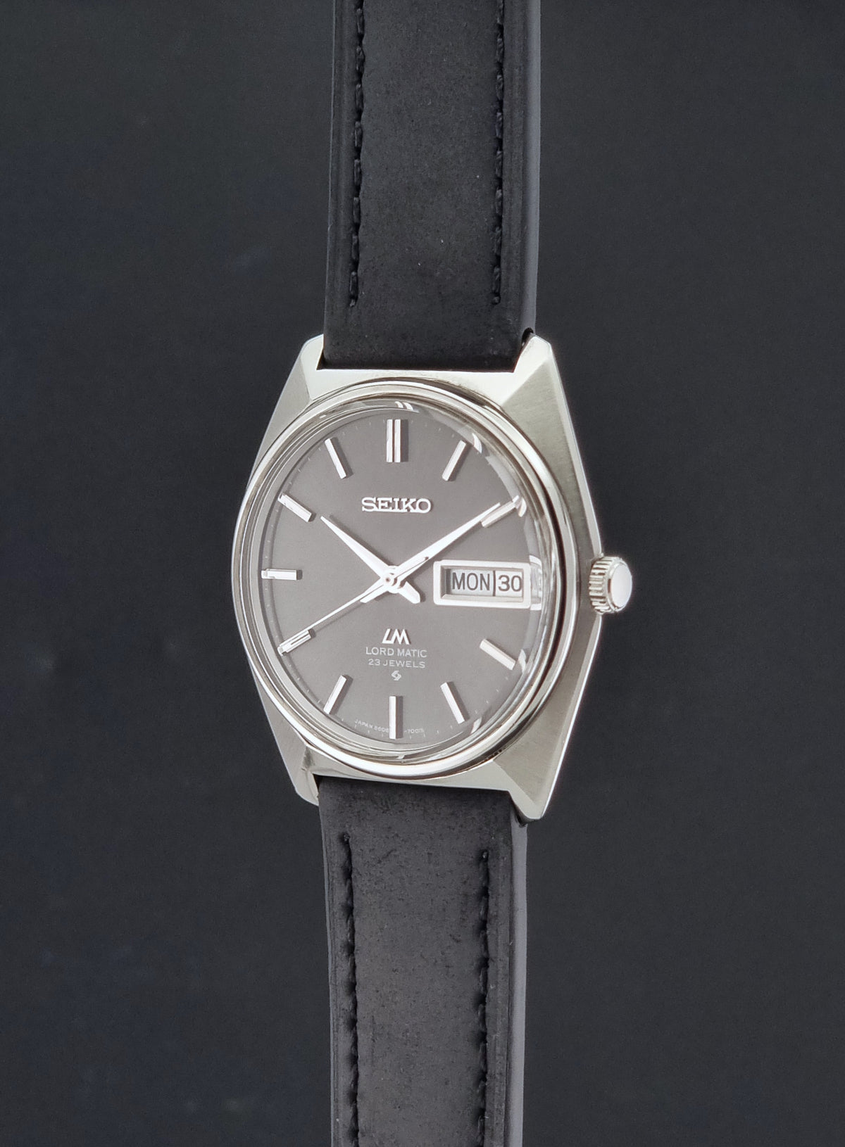 Vintage Seiko watch LordMatic 5606-7000 with a silver-toned metal case, a gray dial, and a black leather strap, shown against a dark background.