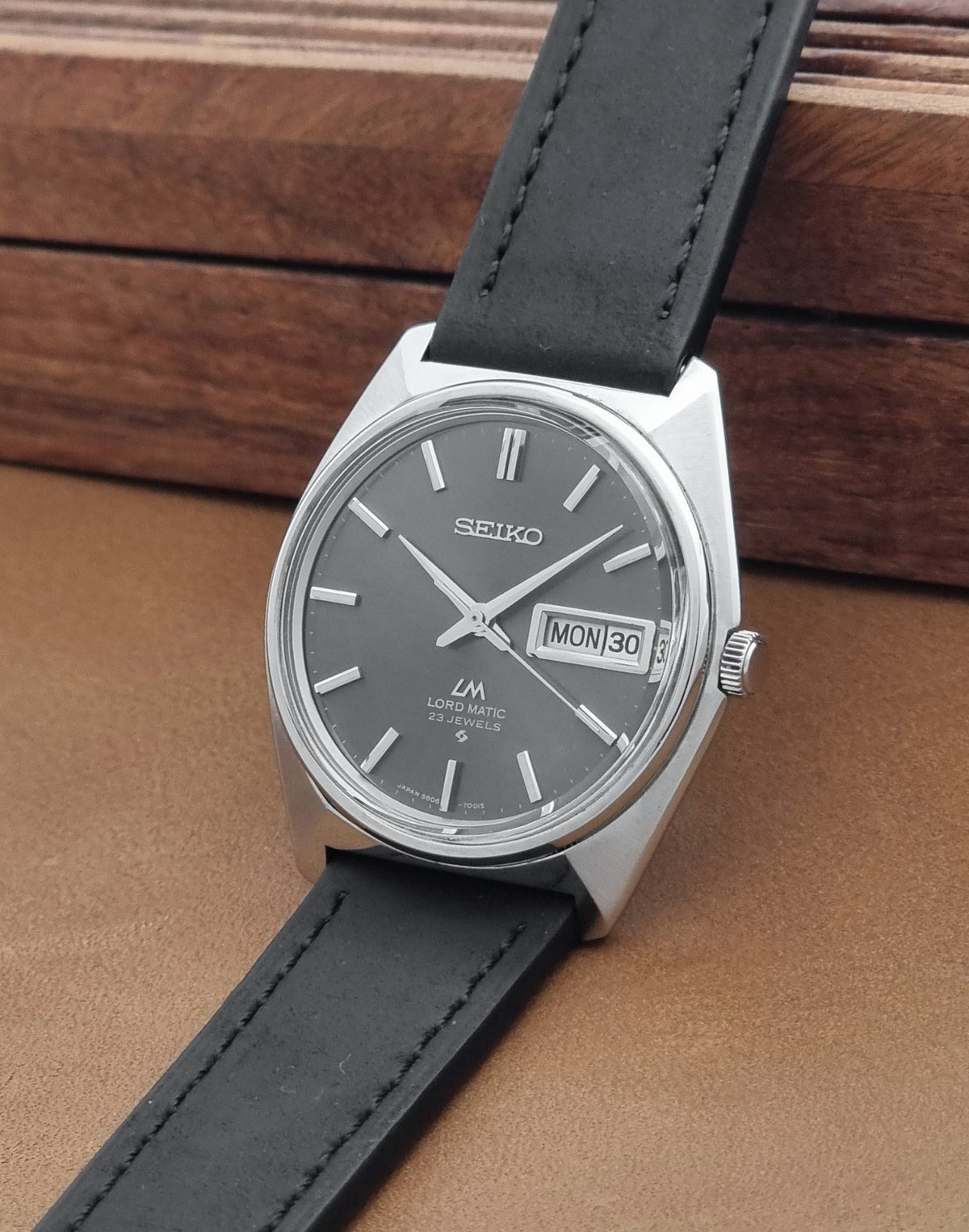 Vintage Seiko watch LordMatic 5606-7000 with a silver-toned metal case, dark gray dial, and black leather strap, resting on a brown leather surface with a wooden background.