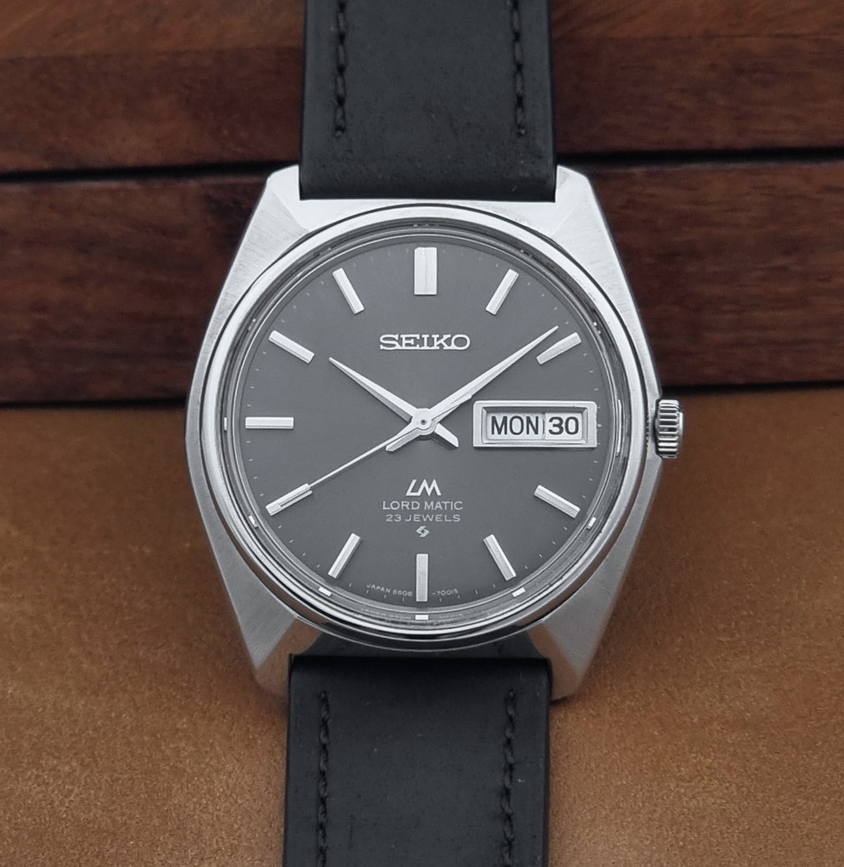 Vintage Seiko watch LordMatic 5606-7000 with a silver-toned metal case, dark gray dial, and black leather strap, photographed on a brown leather surface with a wooden background.