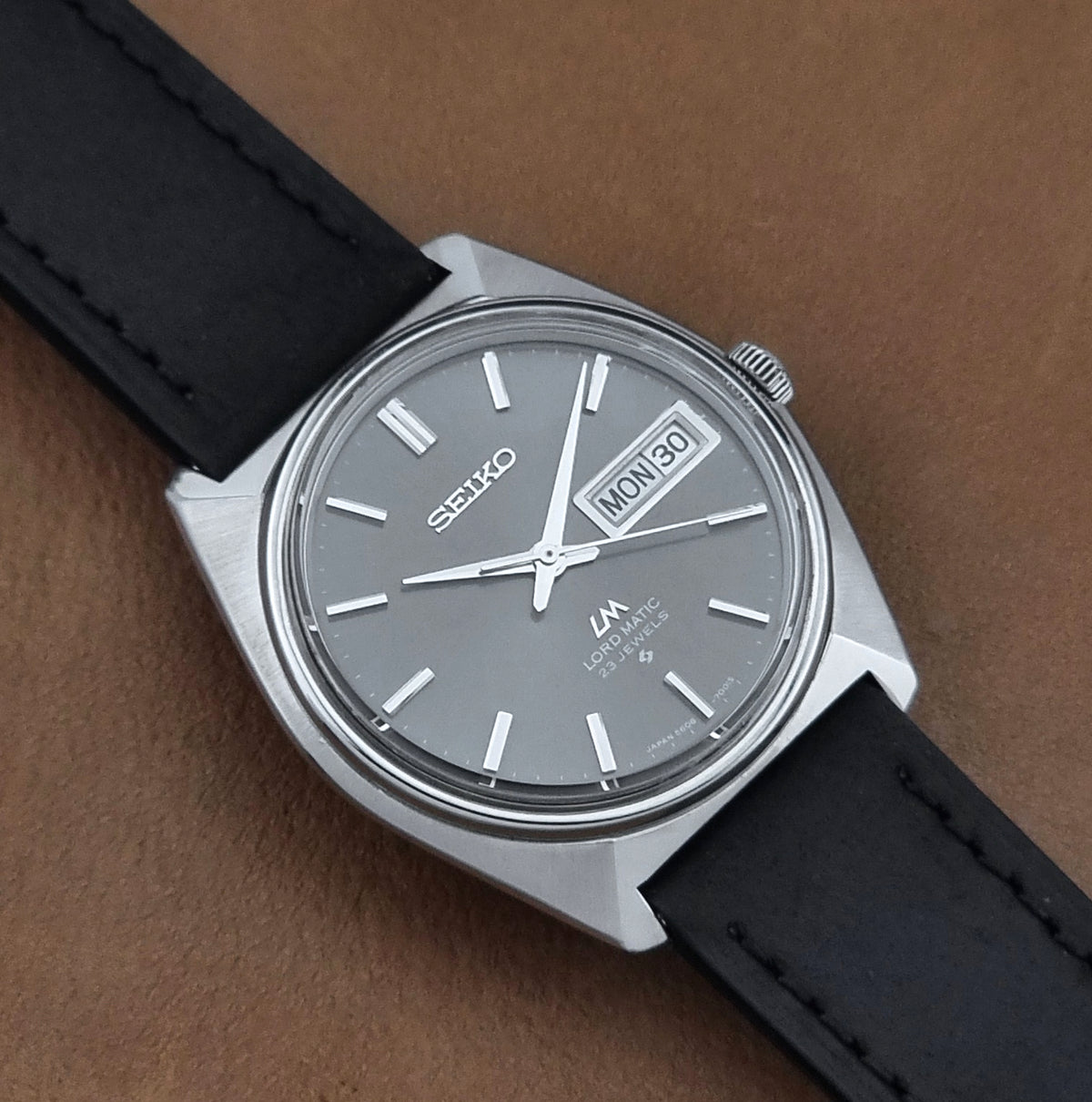 Vintage Seiko watch LordMatic 5606-7000 with a silver-toned metal case, a gray dial, and a black leather strap, photographed on a brown textured surface.