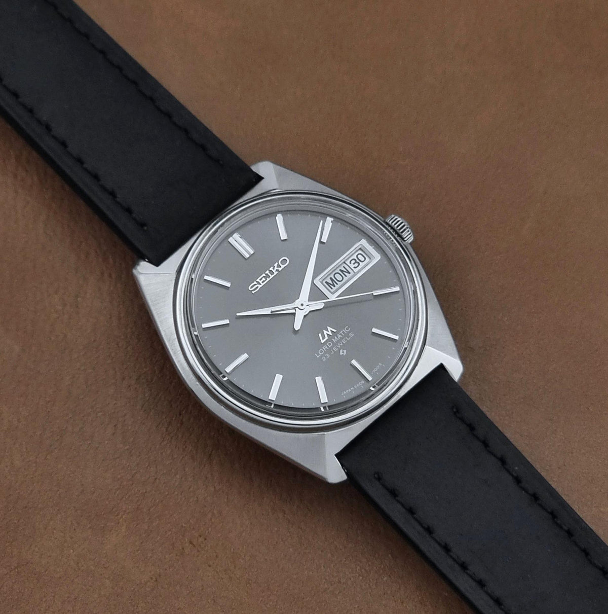 Vintage Seiko watch LordMatic 5606-7000 with a silver-toned metal case, a gray dial, and a black leather strap, photographed diagonally on a brown textured surface.