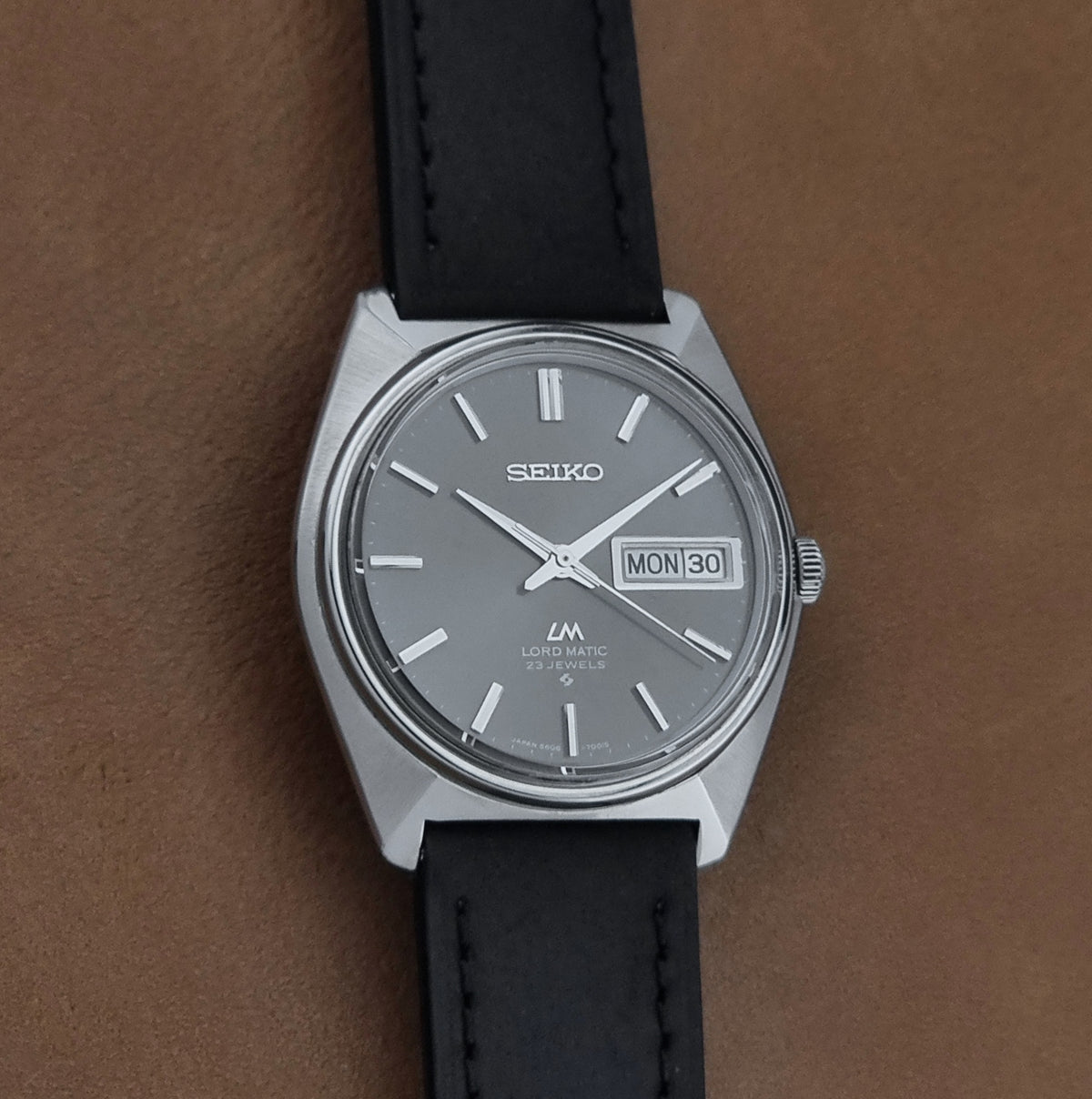 Vintage Seiko watch LordMatic 5606-7000 with a silver-toned metal case, a gray dial, and a black leather strap, shown on a textured brown background.