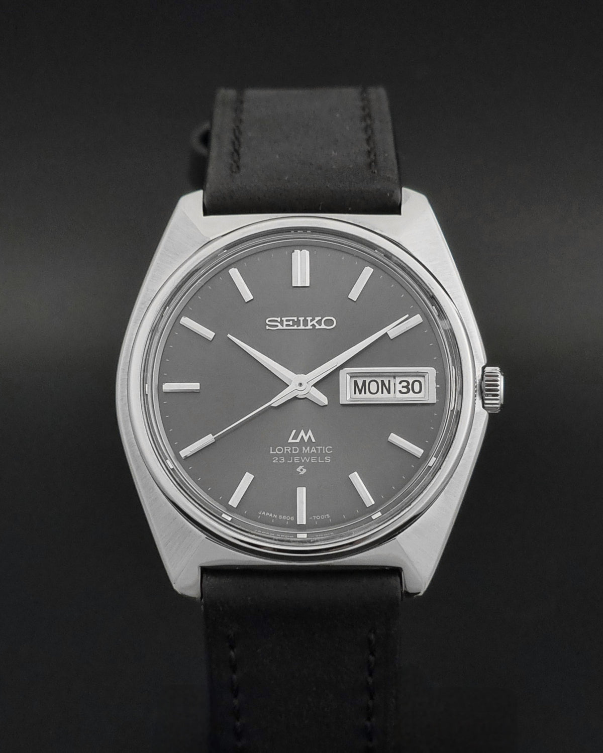 A Seiko LM Automatic 23 Jewels wristwatch with a silver-tone case, dark gray dial, and black leather strap, shown against a dark background.