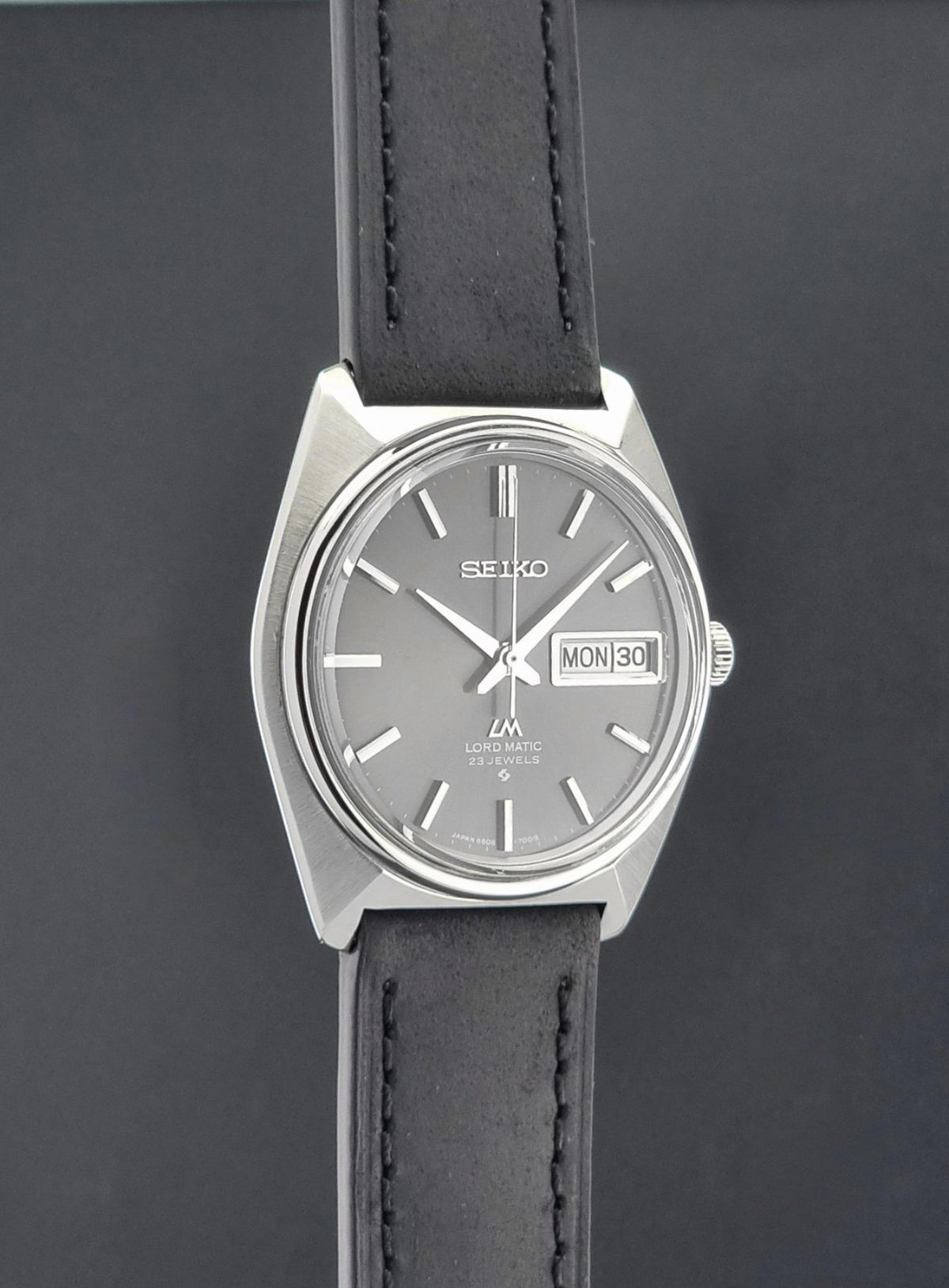 Vintage Seiko watch LordMatic 5606-7000 with a silver-toned metal case, a gray dial, and a black leather strap, shown against a dark gray background.