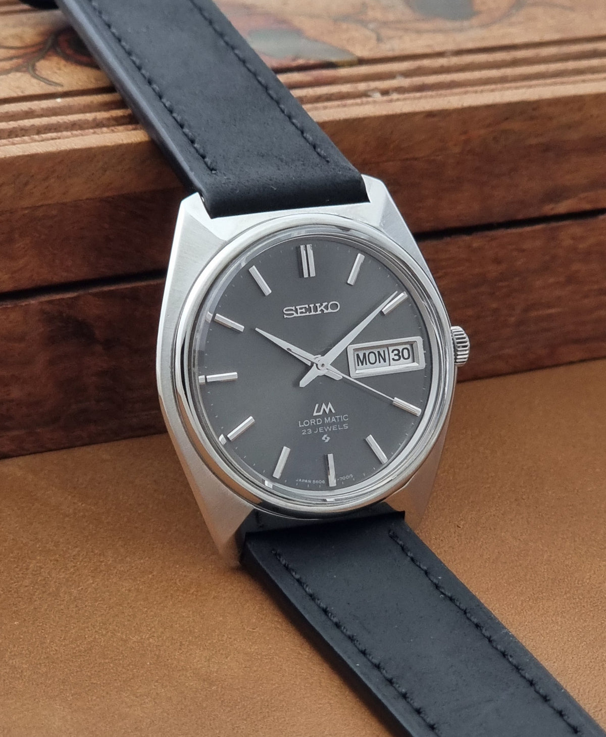 Vintage Seiko watch LordMatic 5606-7000 with a silver-toned metal case, dark gray dial, and black leather strap, resting on a brown wooden surface.