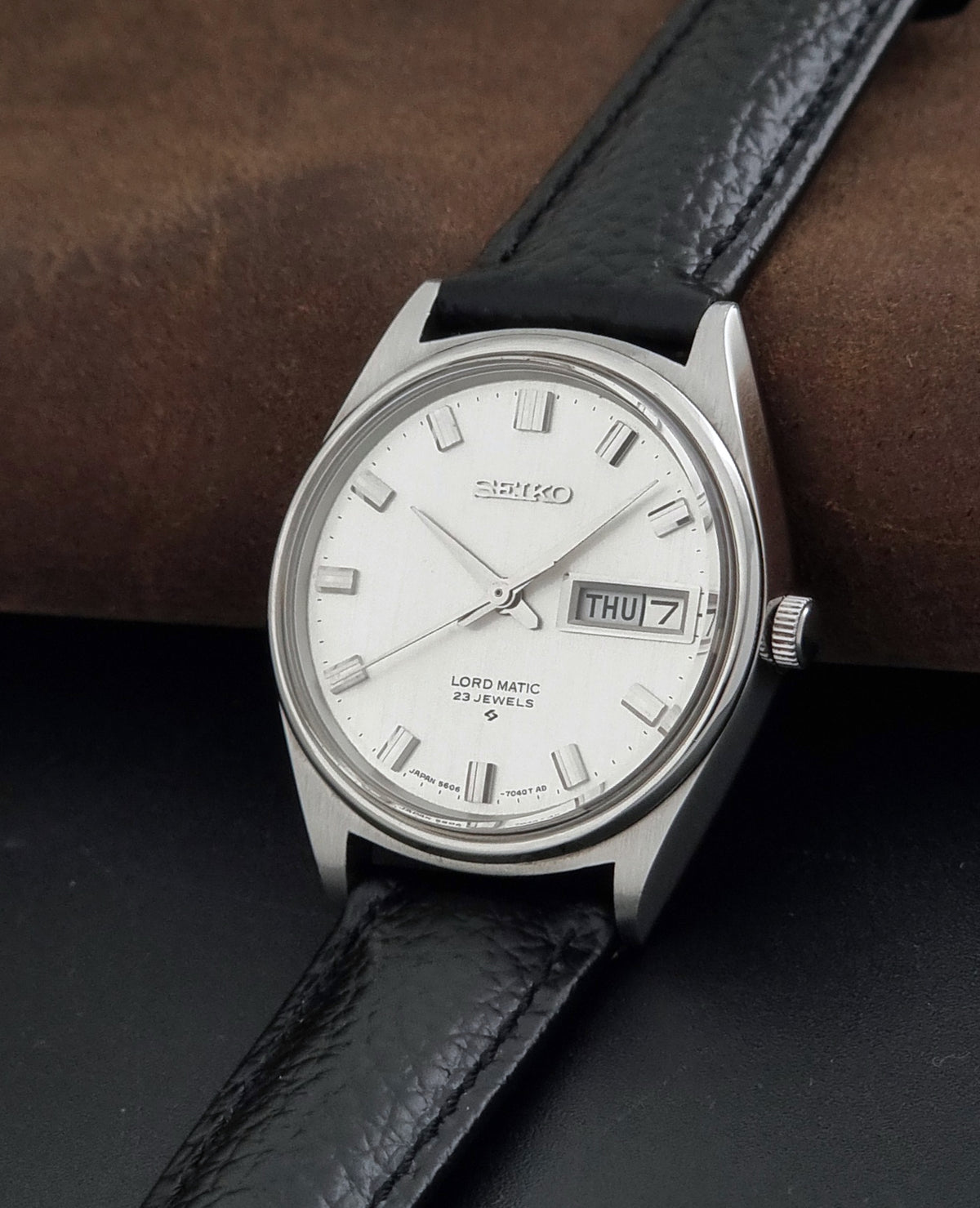 A Vintage Seiko watch LordMatic 5606-7040 with a silver-toned metal case, white dial, and black leather strap, resting on a dark surface with a brown textured background.