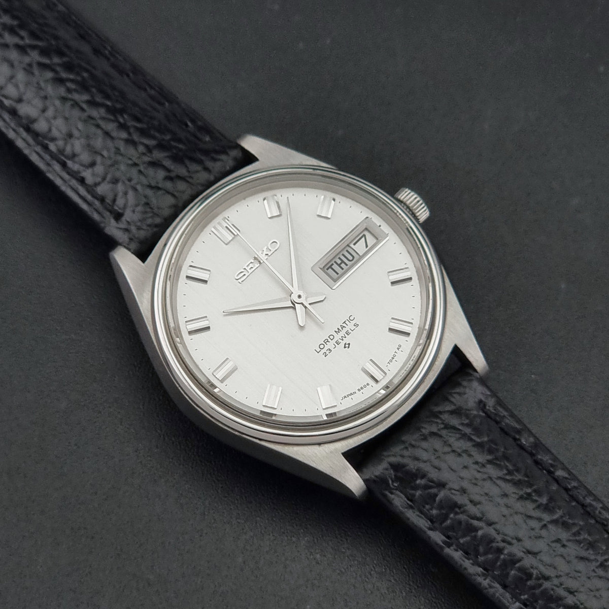 A Vintage Seiko watch LordMatic 5606-7040 with a silver-toned metal case, white dial, and black textured leather strap, photographed on a dark gray background.