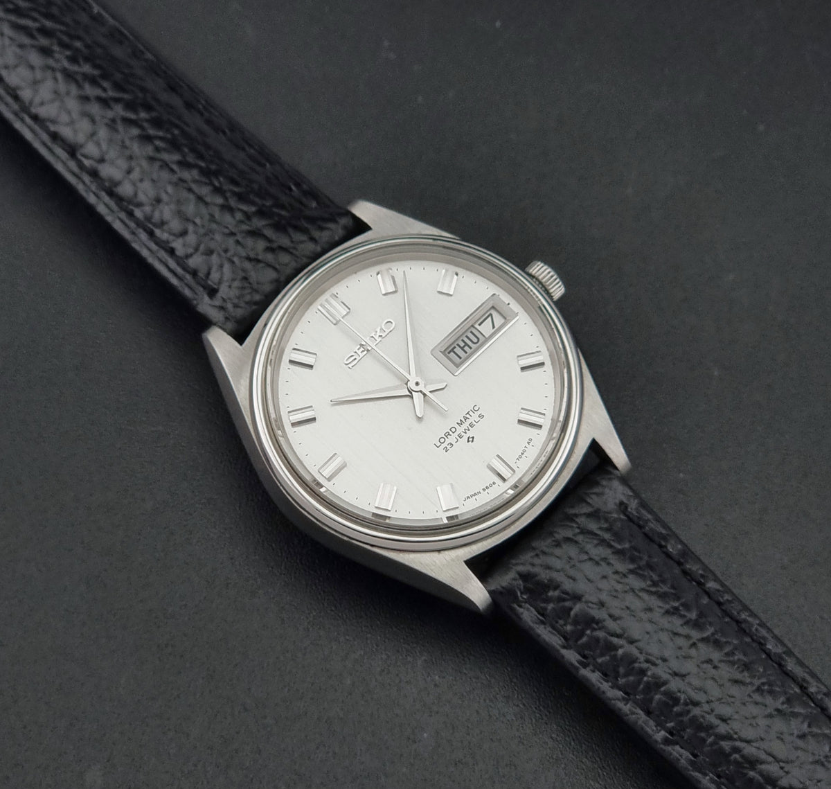 A Vintage Seiko watch LordMatic 5606-7040 with a silver-toned metal case, white dial, and black textured leather strap, photographed diagonally on a dark gray background.