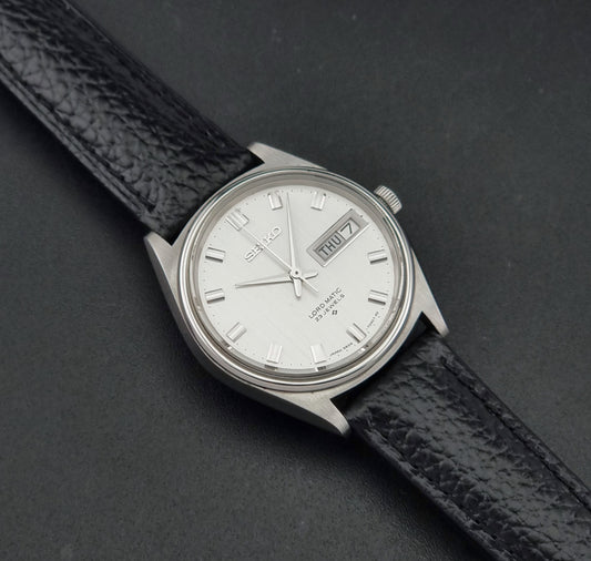 A Vintage Seiko watch LordMatic 5606-7040 with a silver-toned metal case, white dial, and black textured leather strap, photographed diagonally on a dark gray background.