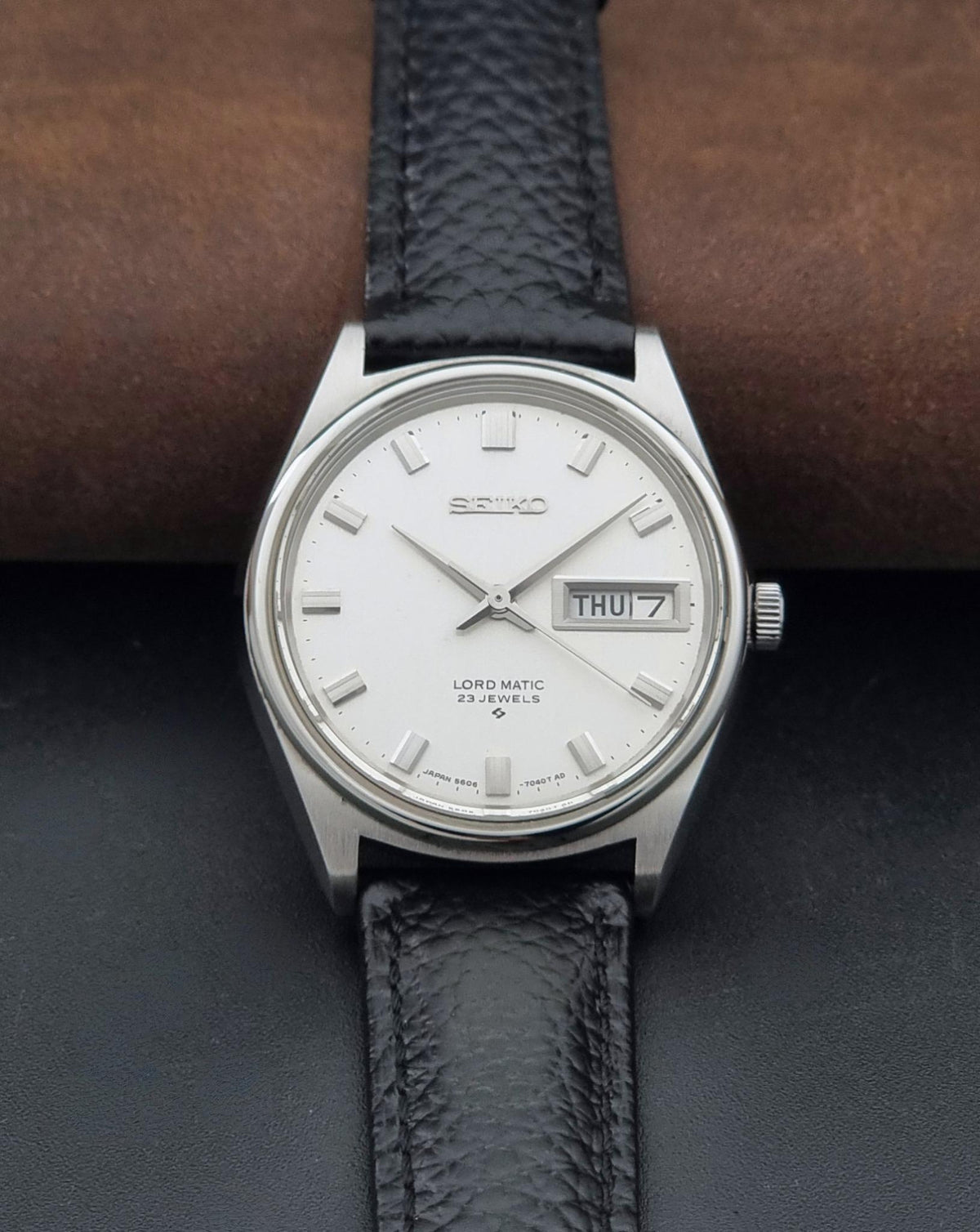 A Vintage Seiko watch LordMatic 5606-7040 with a silver-toned metal case, white dial, and textured black leather strap, resting on a dark surface with a brown cylindrical prop behind it.